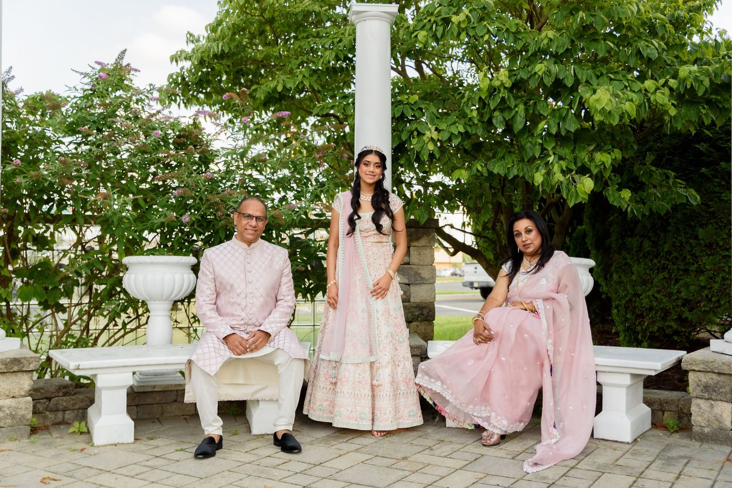 A wedding party in soft pink dresses poses together on white steps surrounded by lush green trees.