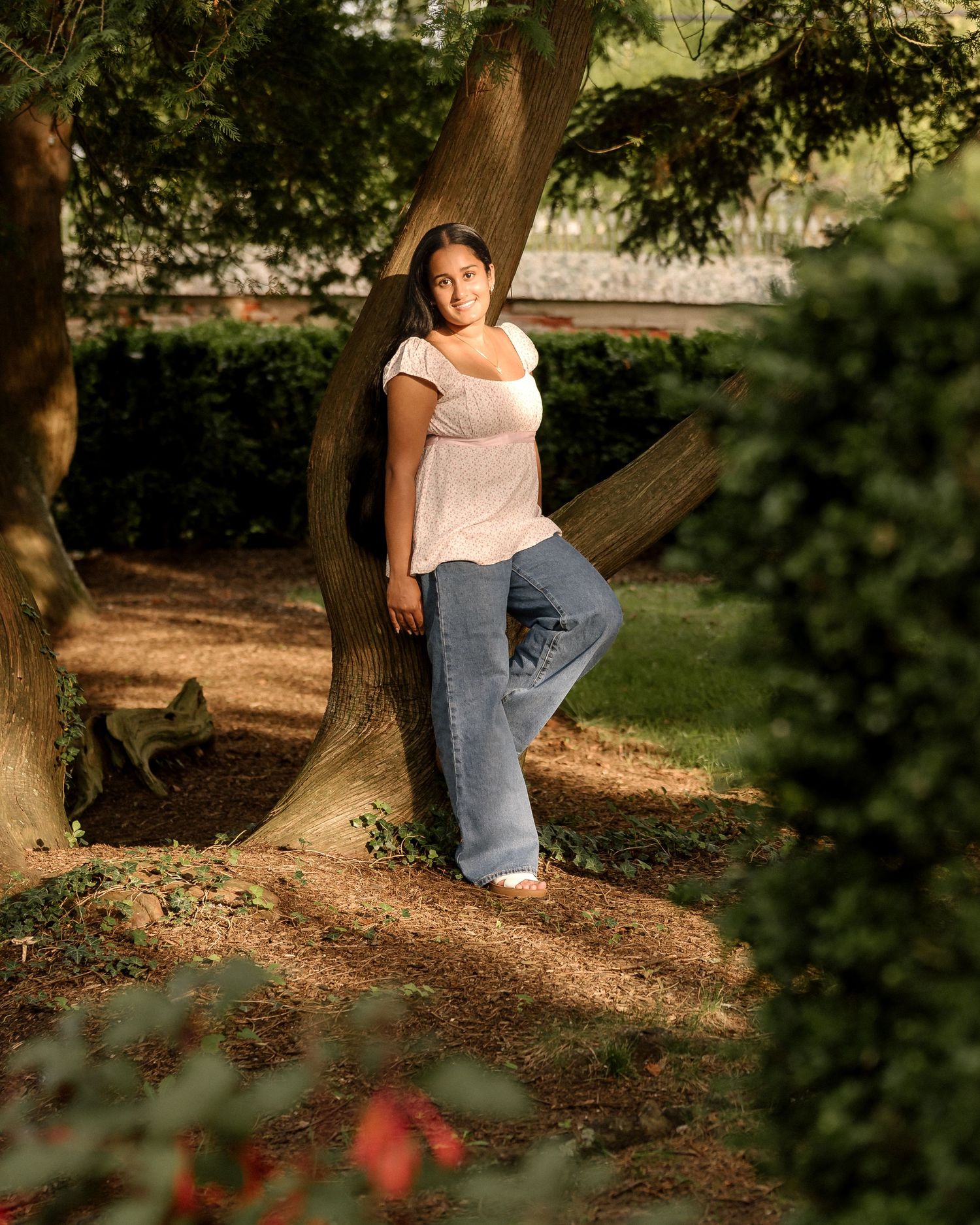 A person sits in dappled sunlight on a wooded path wearing jeans and a light colored top in a natural setting.