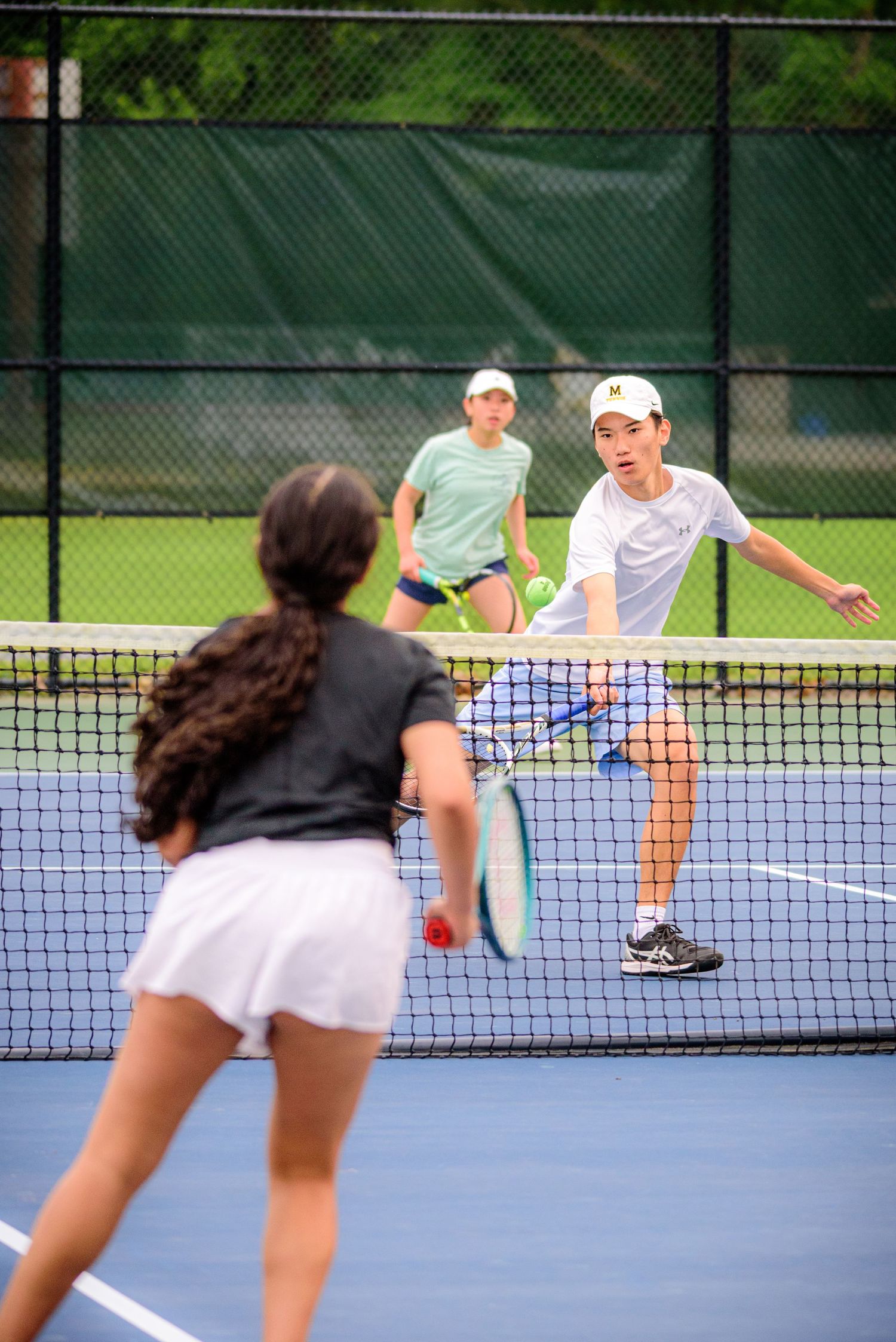 Tennis players practicing volleys at the net on an outdoor blue court with chain link fencing in the background.