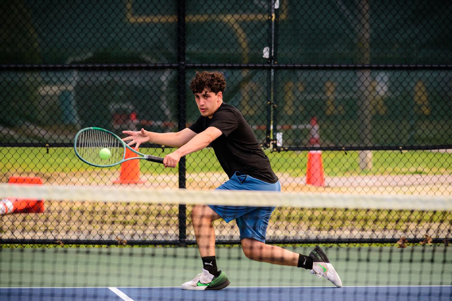 Tennis player in black shirt and blue shorts stretches to make a forehand return on outdoor court.