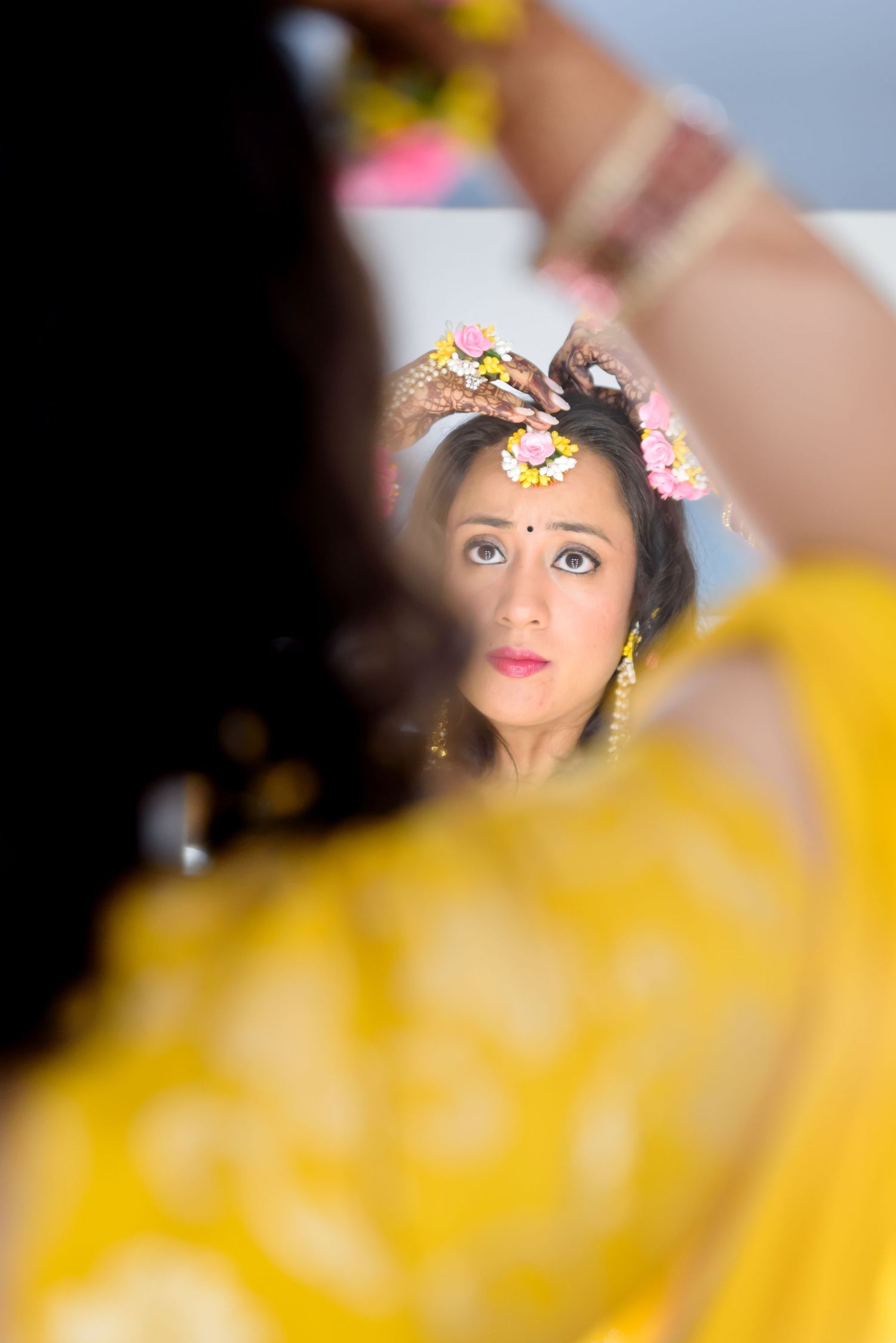 Indian bride in yellow saree with floral hair accessories checks her look in mirror during wedding preparation.