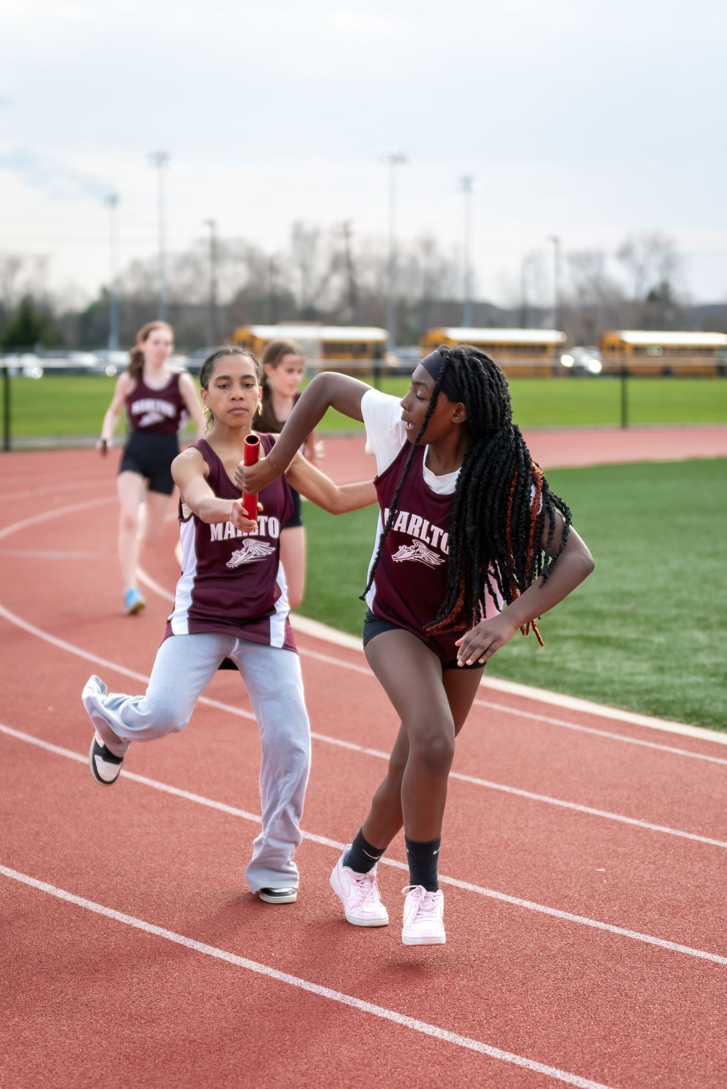 Two female track athletes in maroon uniforms exchange a baton during a relay race on a red track.