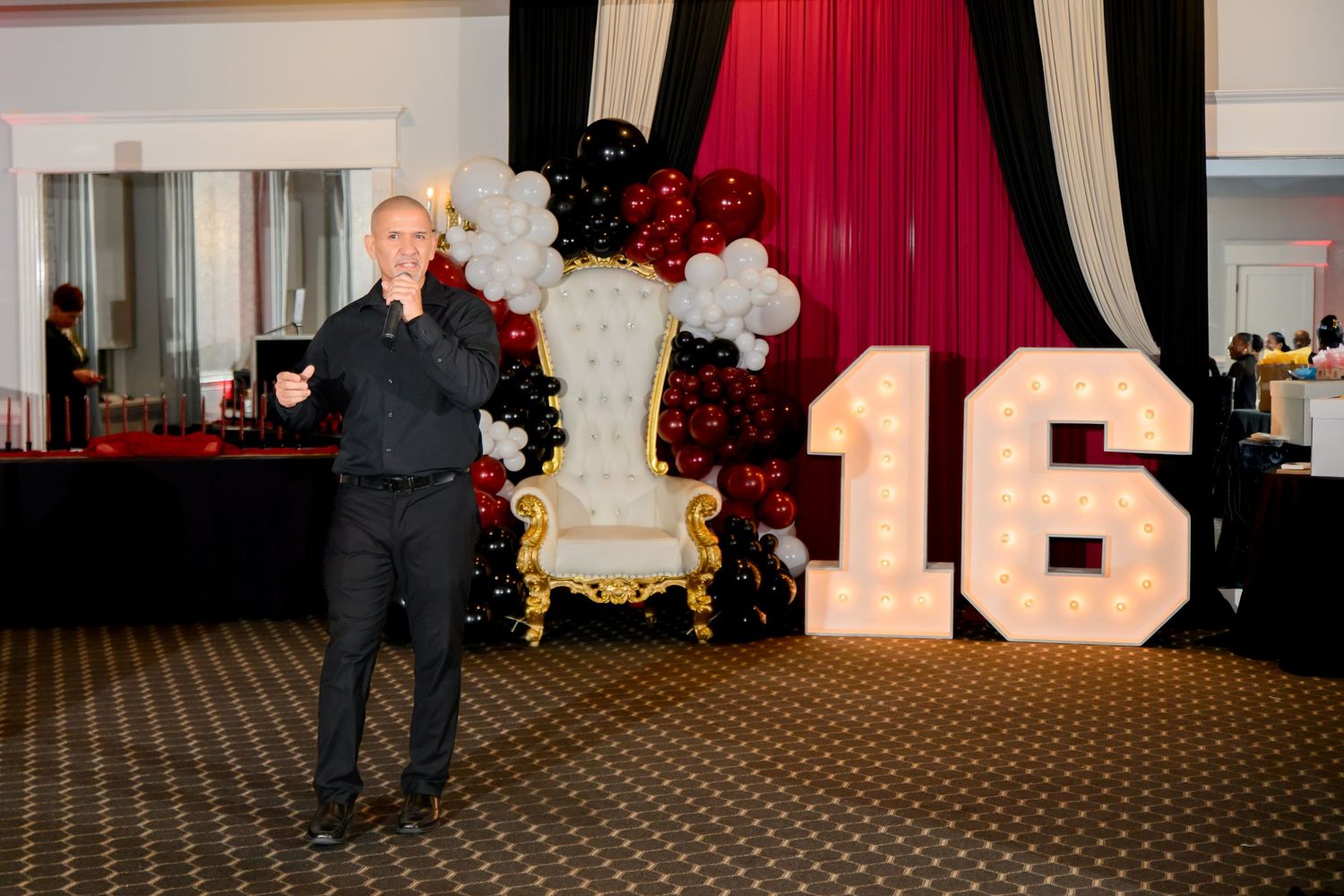 Man posing at a Sweet 16 party with marquee numbers, gold throne chair, and black and red balloon decor.