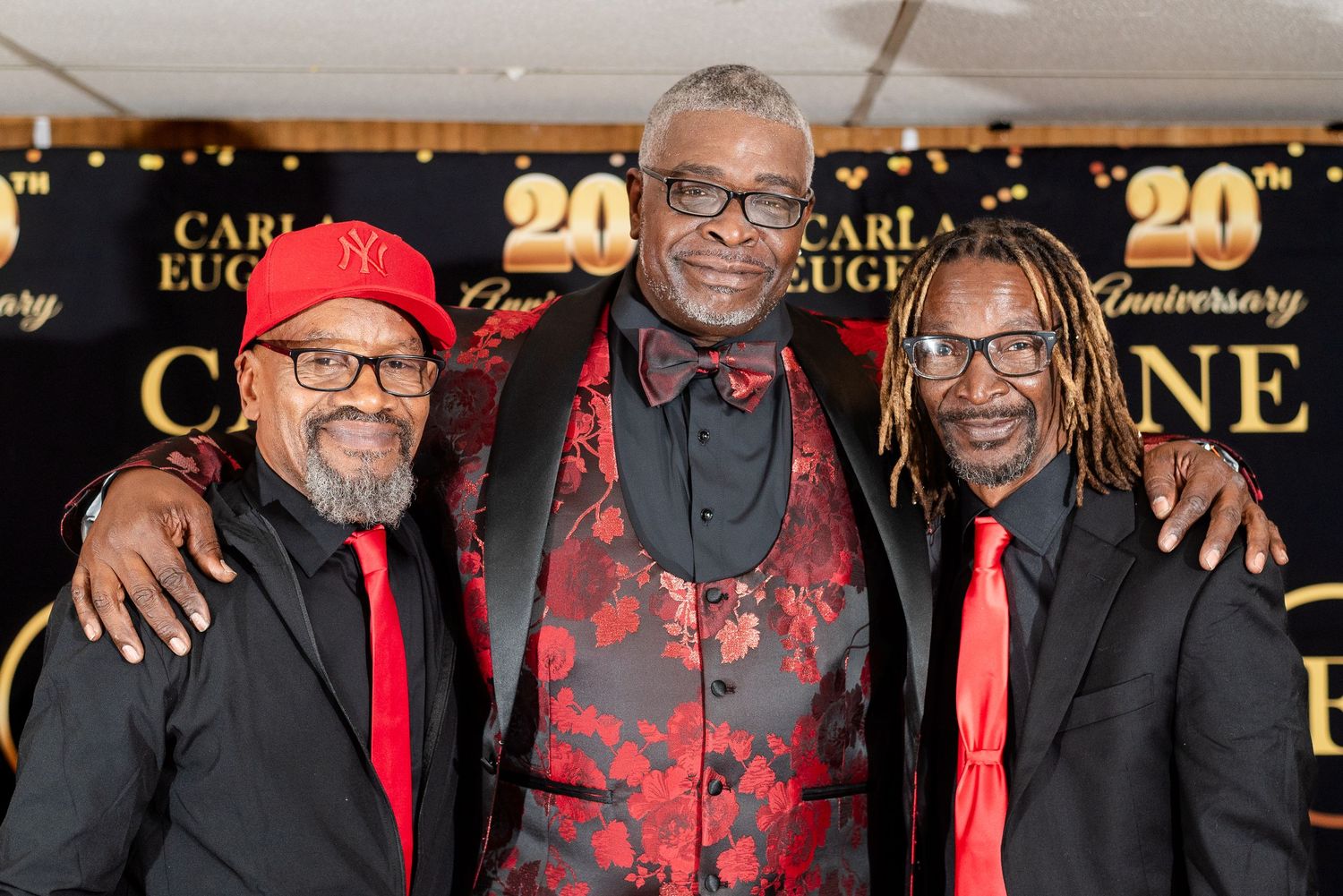 Three men in red and black formal attire smile together at a 20th anniversary celebration event.