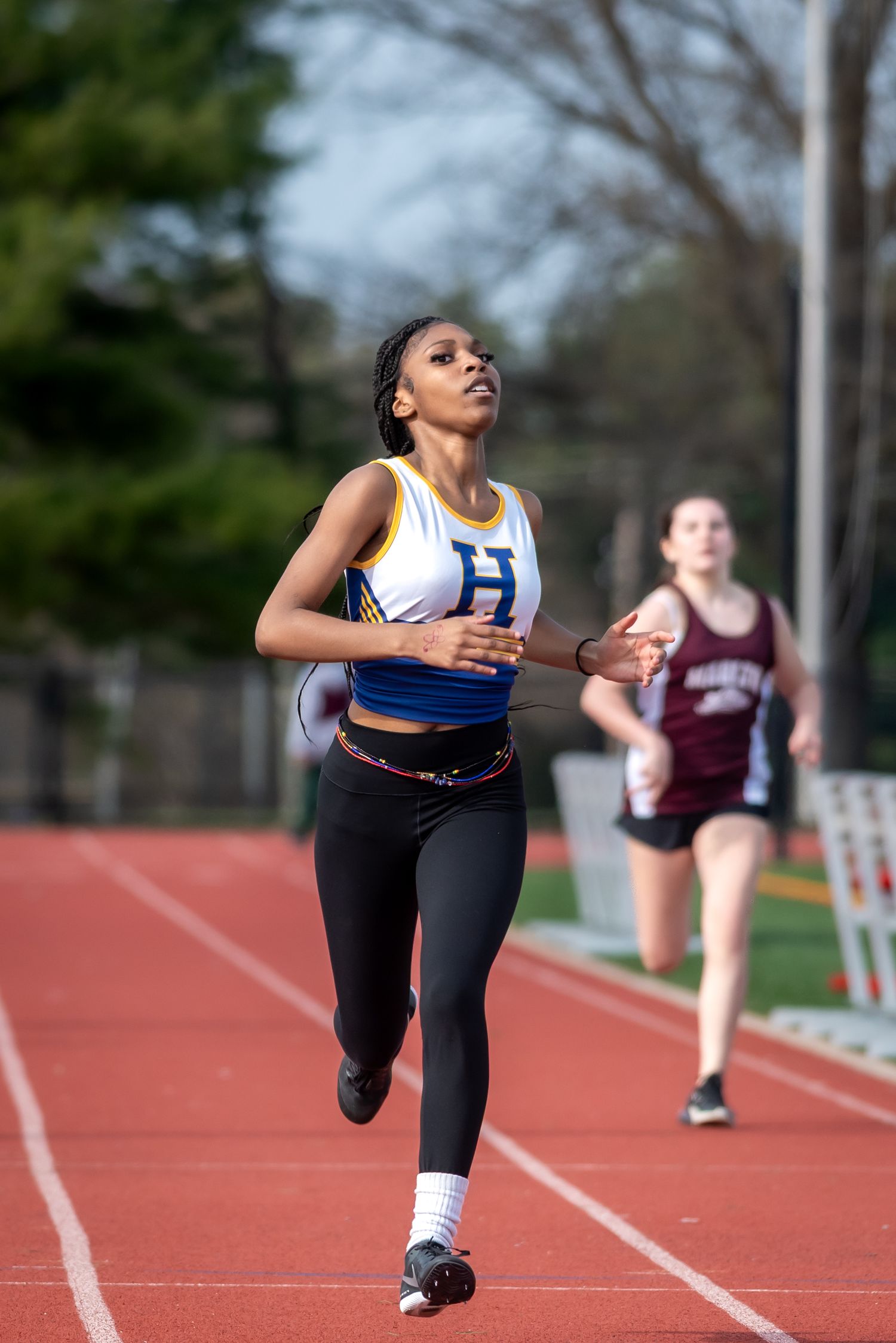 Two female athletes competing in a track race, with the lead runner wearing a blue and gold H jersey.