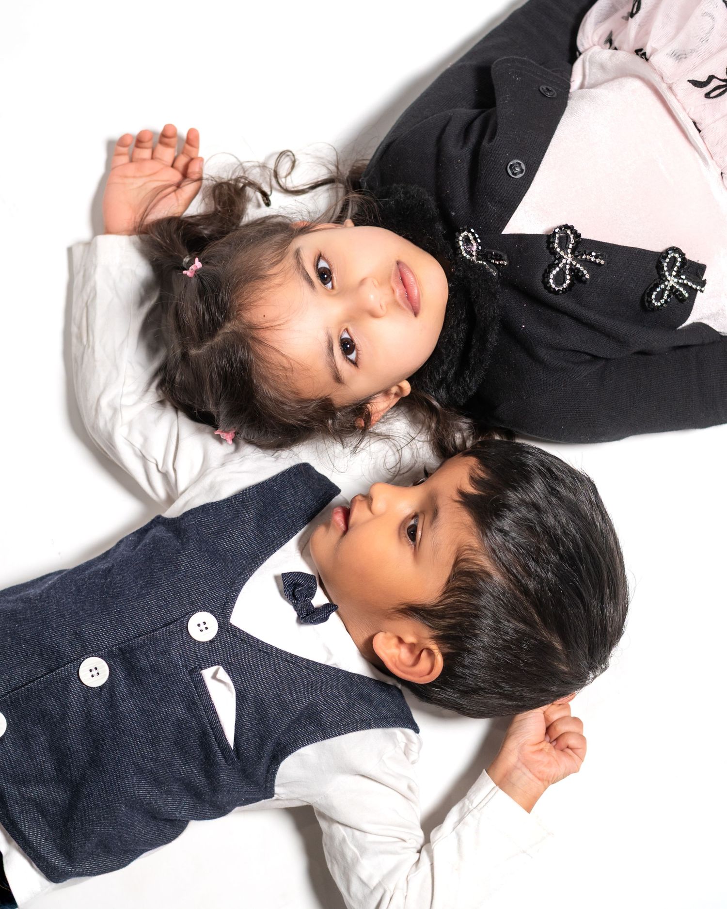 Two young children lying down together, a girl in a black top and a boy in a navy vest with bow tie.