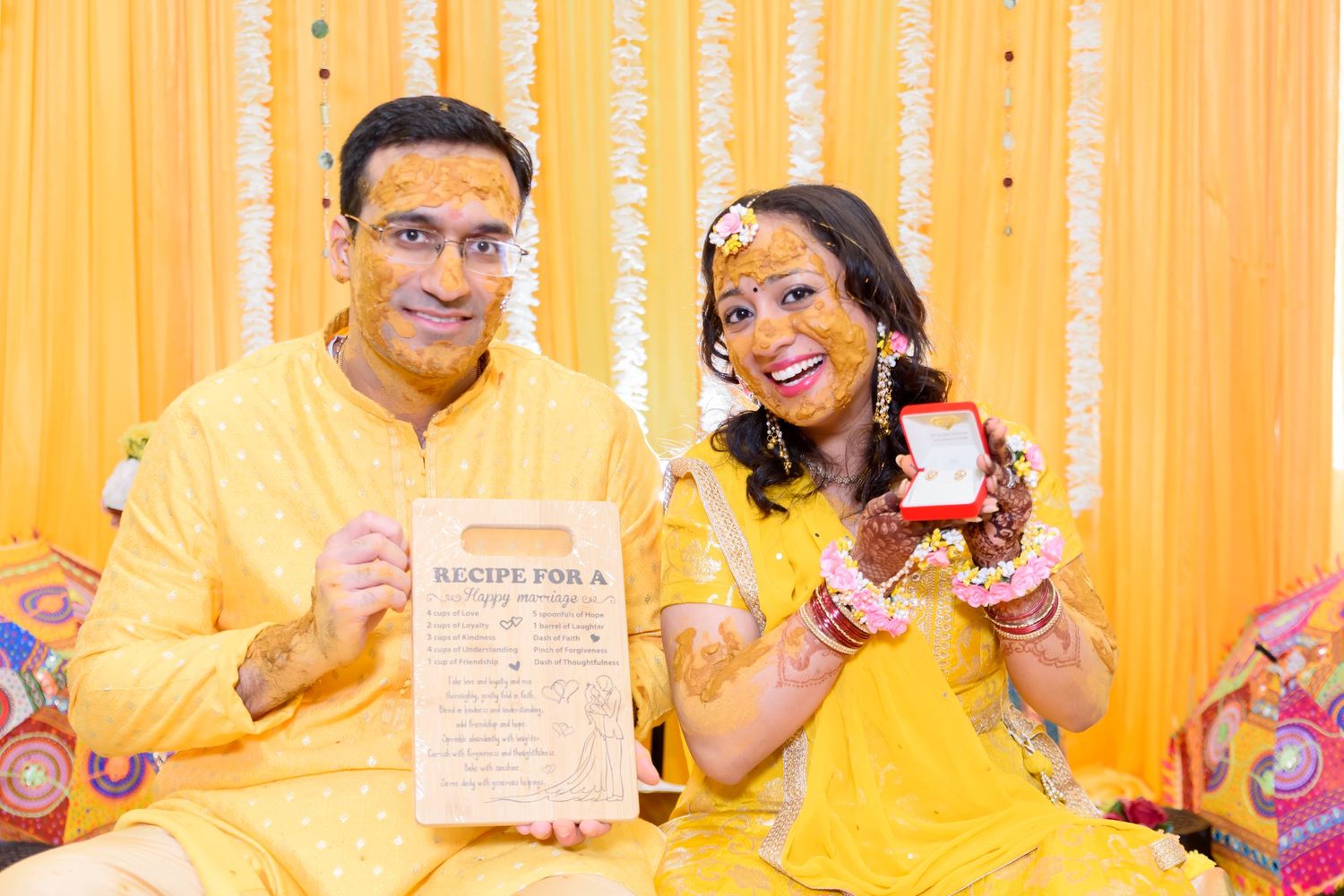 Indian couple in yellow outfits at Haldi ceremony holding ring box and recipe card against orange backdrop.