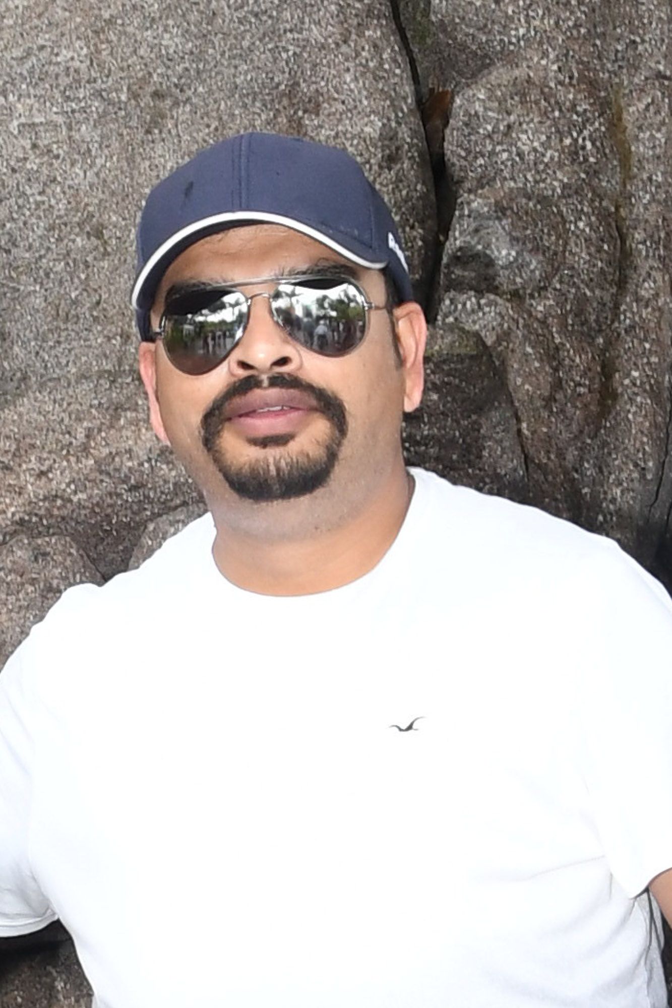 Man wearing blue cap and aviator sunglasses in white t-shirt posing outdoors near rocky background.