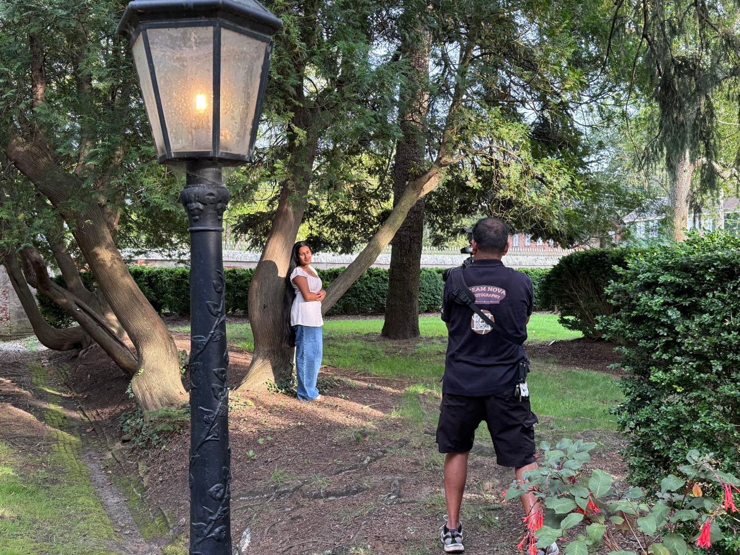 People taking photos in a garden setting with a vintage-style lamppost and lush greenery.