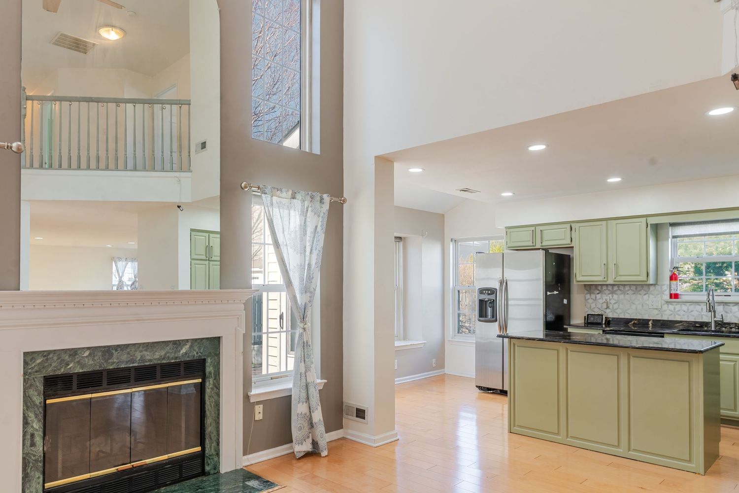 Open-concept kitchen with green cabinets, marble fireplace, high ceilings, and hardwood floors.