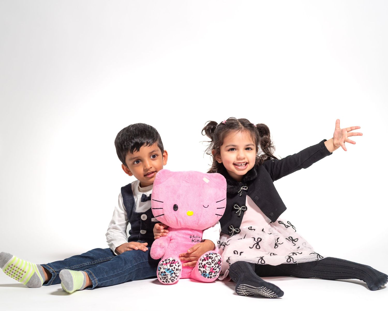 Two young children sit together smiling, holding a pink Hello Kitty plush toy, against a white background.