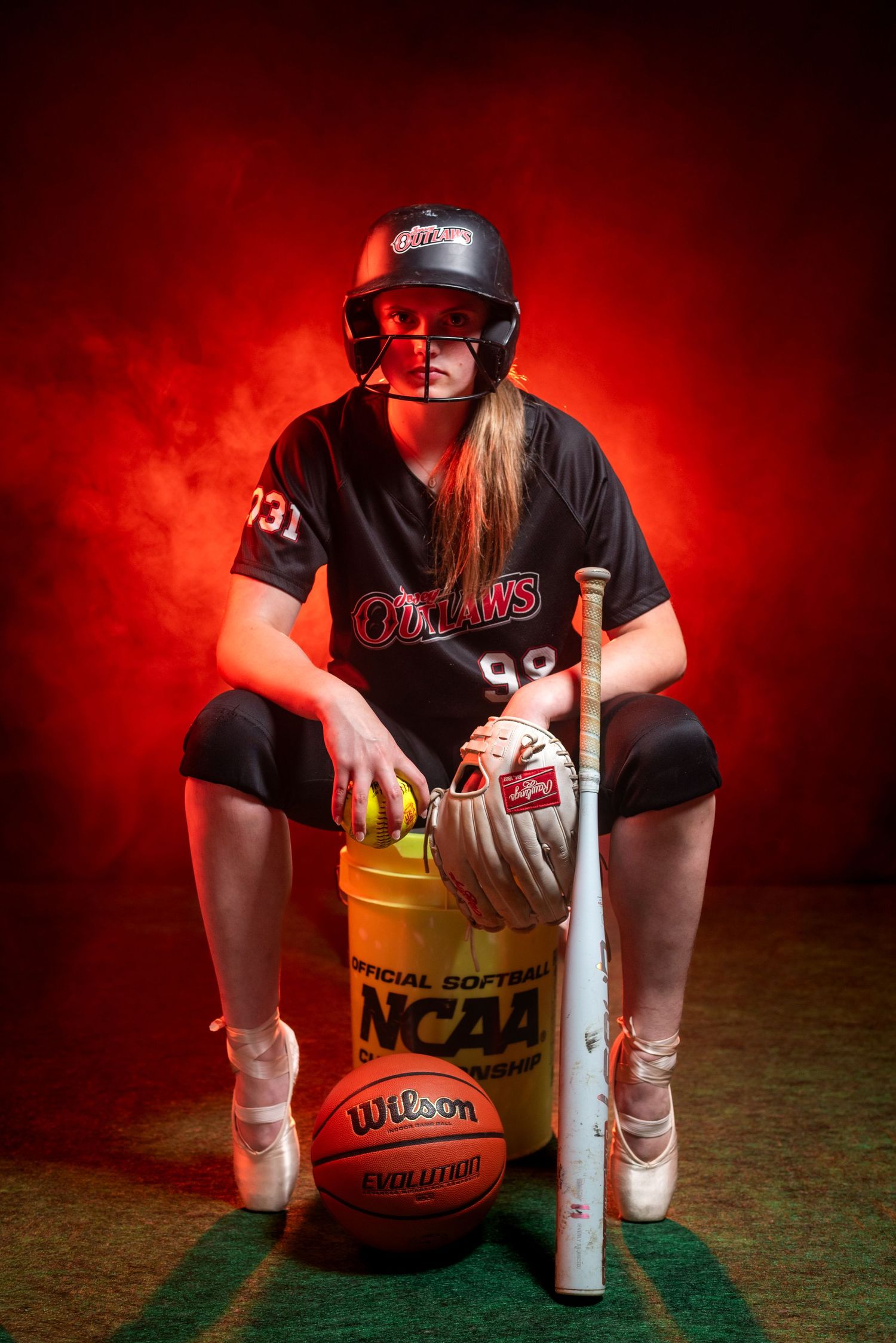 Female softball player in Outlaws uniform poses with bat, glove, and NCAA Wilson softball equipment against red smoky background.