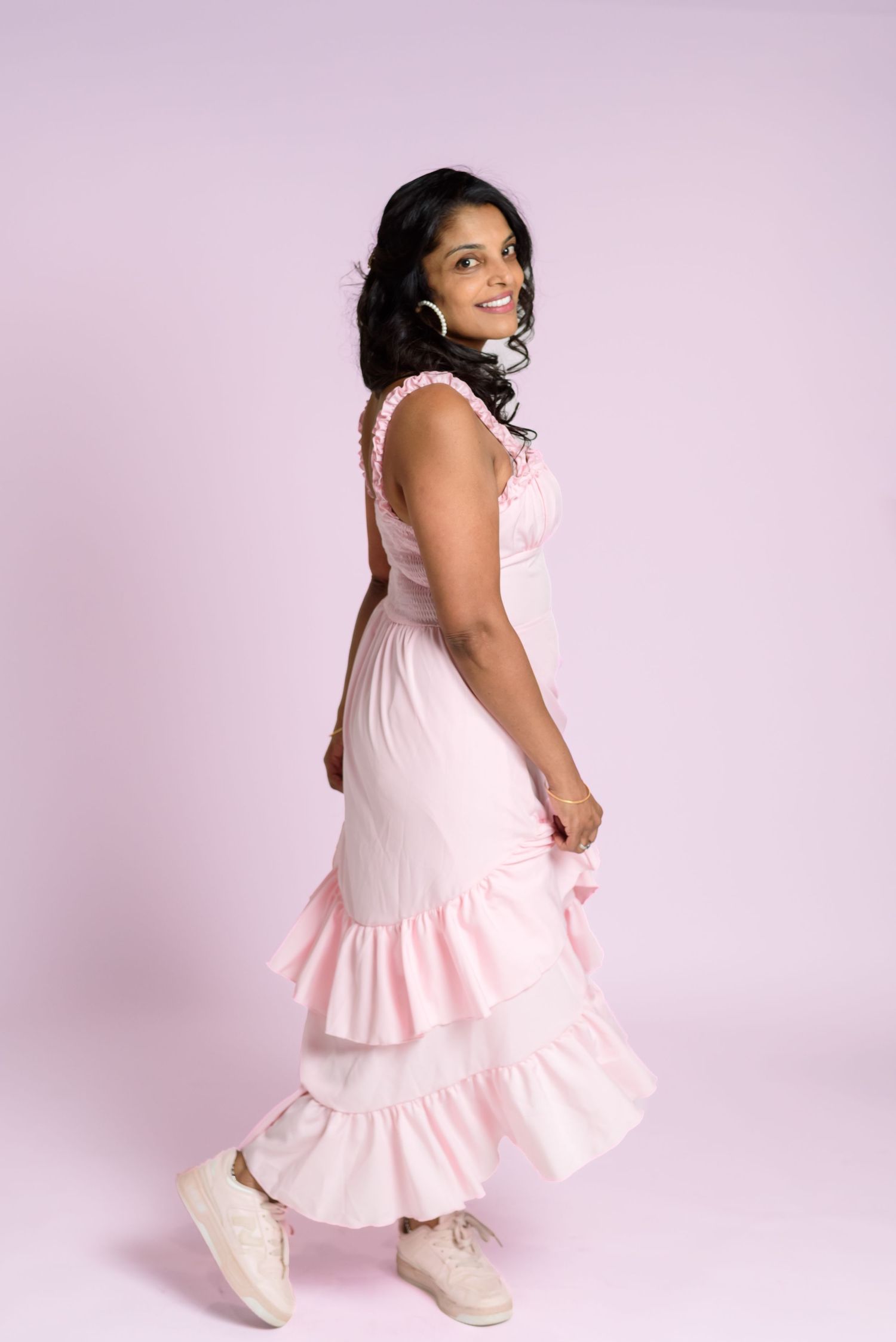 A woman in a pink ruffled midi dress and white sneakers poses gracefully against a soft pink background.