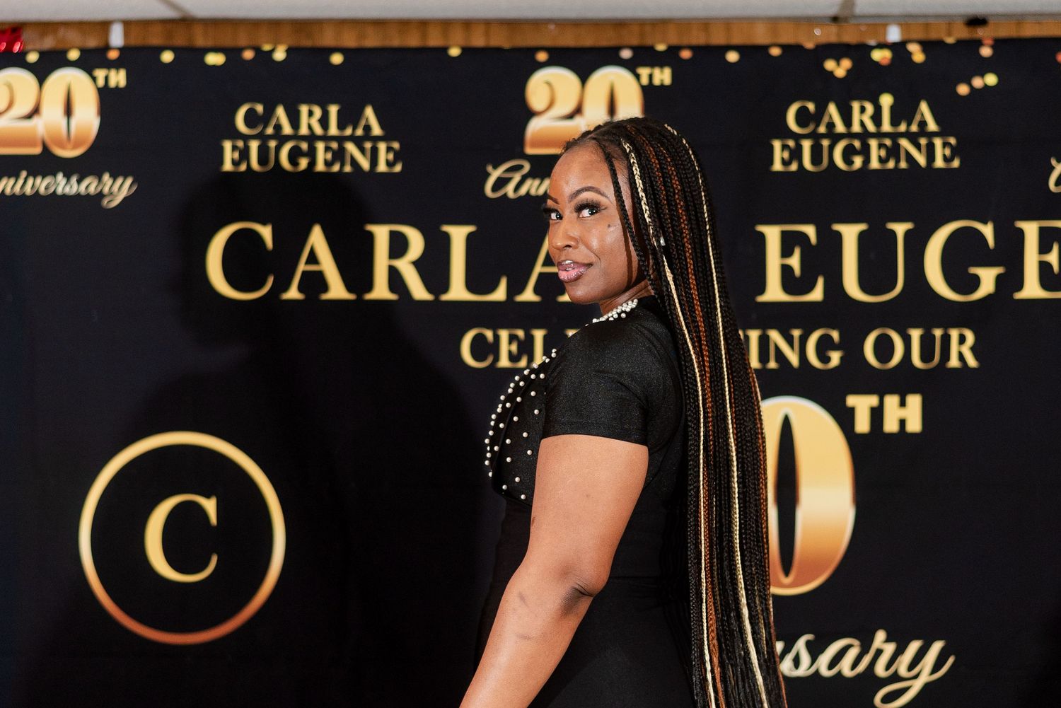 Woman posing at Carla Eugene 20th Anniversary celebration event backdrop in black dress with gold accents.