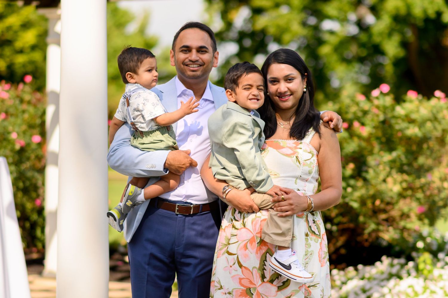 A smiling South Asian family of four poses outdoors, with a man holding a toddler and a woman embracing a young boy.