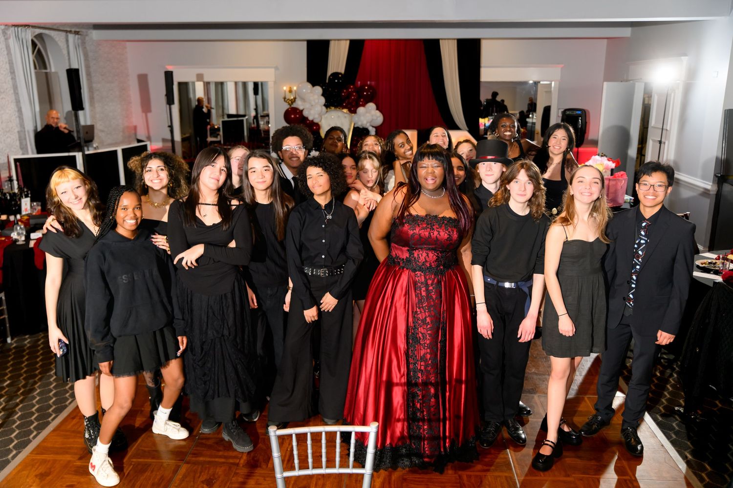 A large group of people dressed in black surround a woman in a red gown at an elegant event.