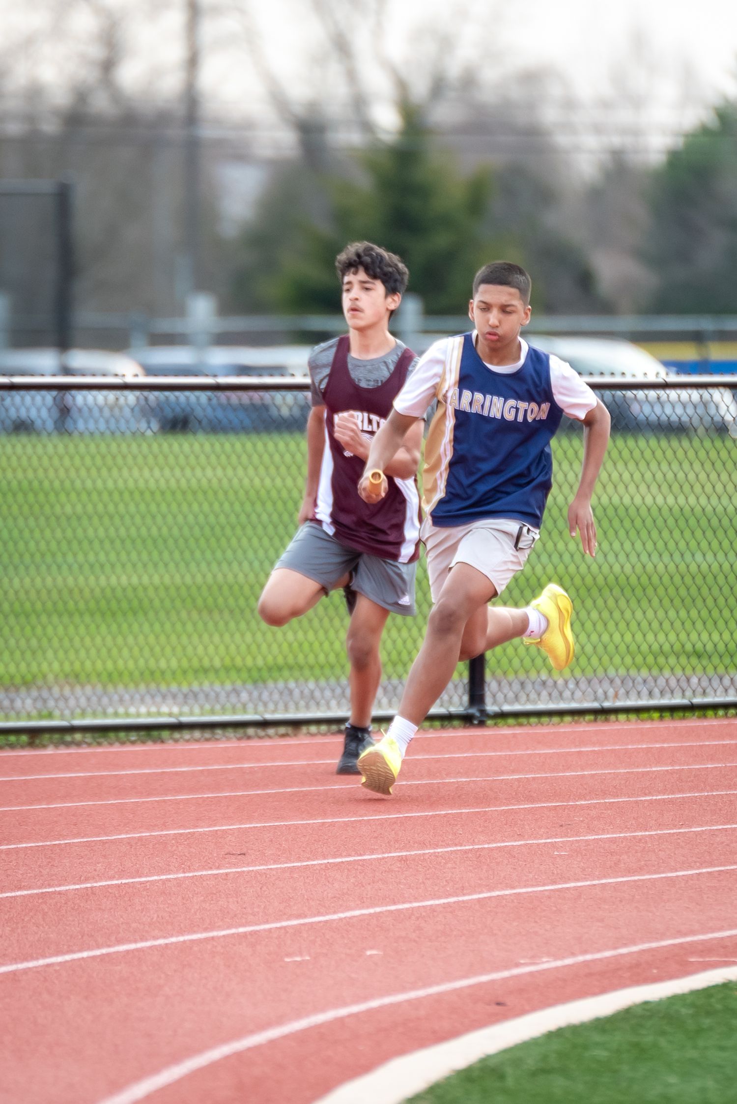 Two young male athletes compete in a track race, wearing Morristown and Barrington jerseys on a red running track.