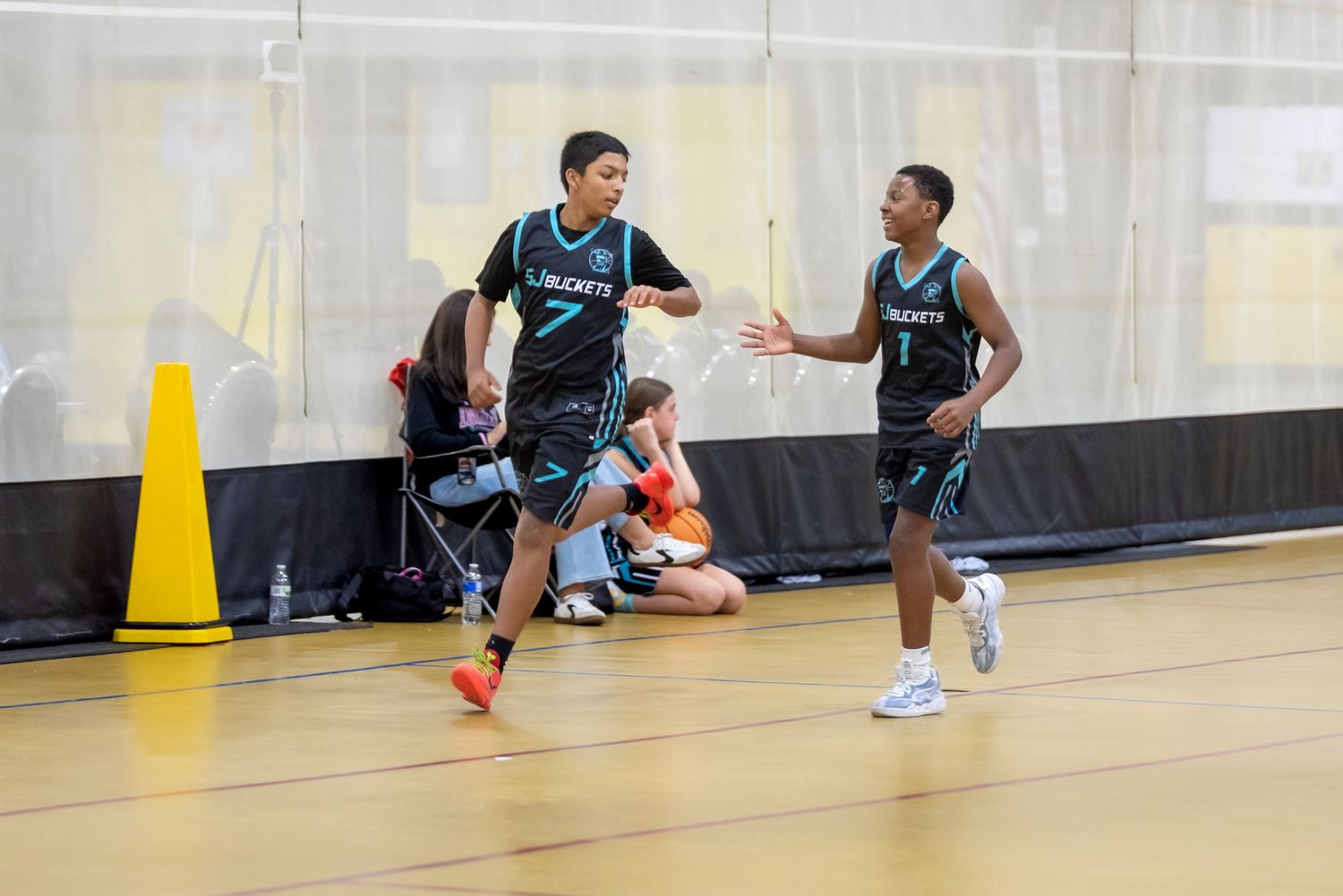 Two young basketball players in black and teal uniforms high-fiving during an indoor basketball game.