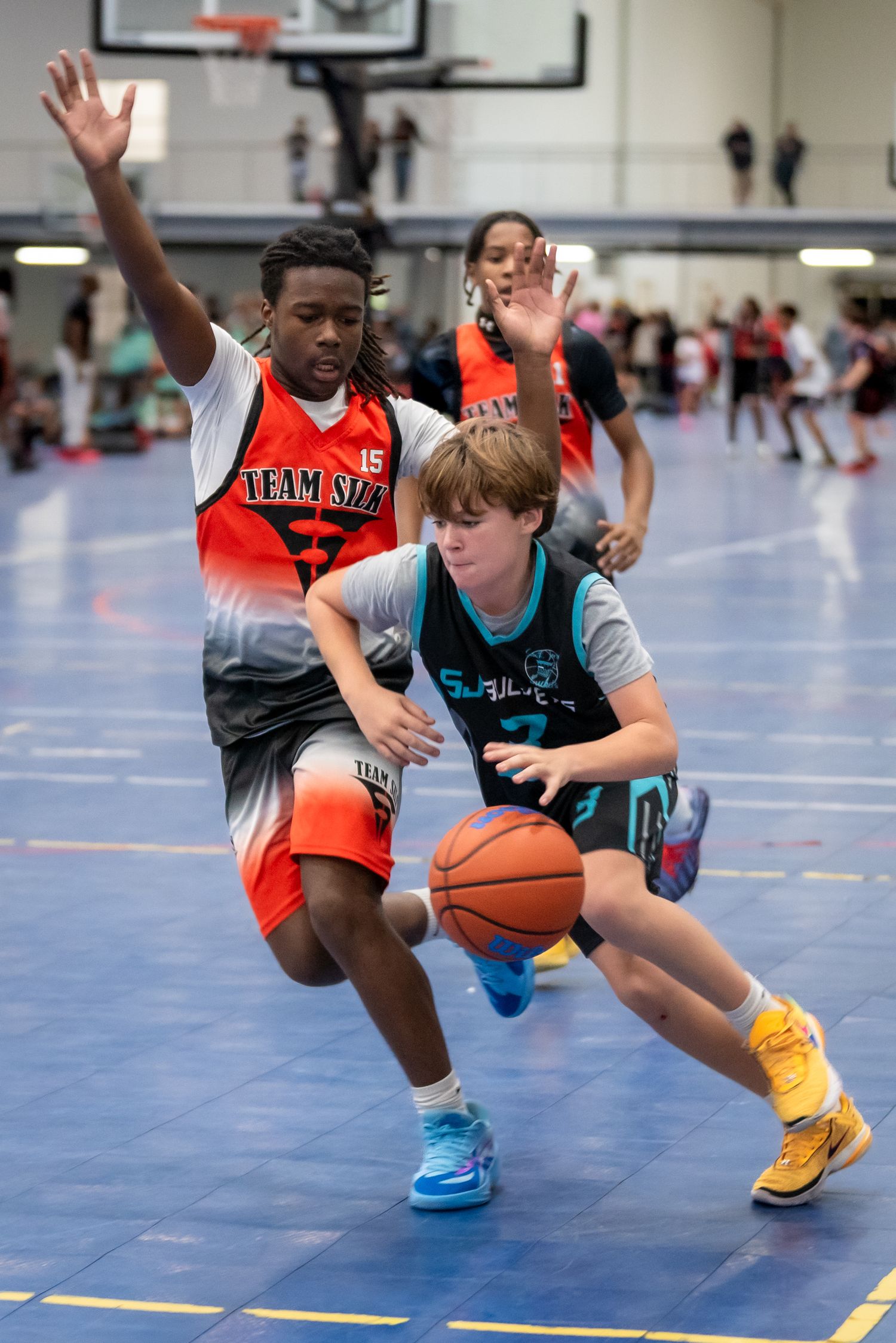 Young basketball players compete in an indoor youth tournament, with one player dribbling past a defender in orange jersey.