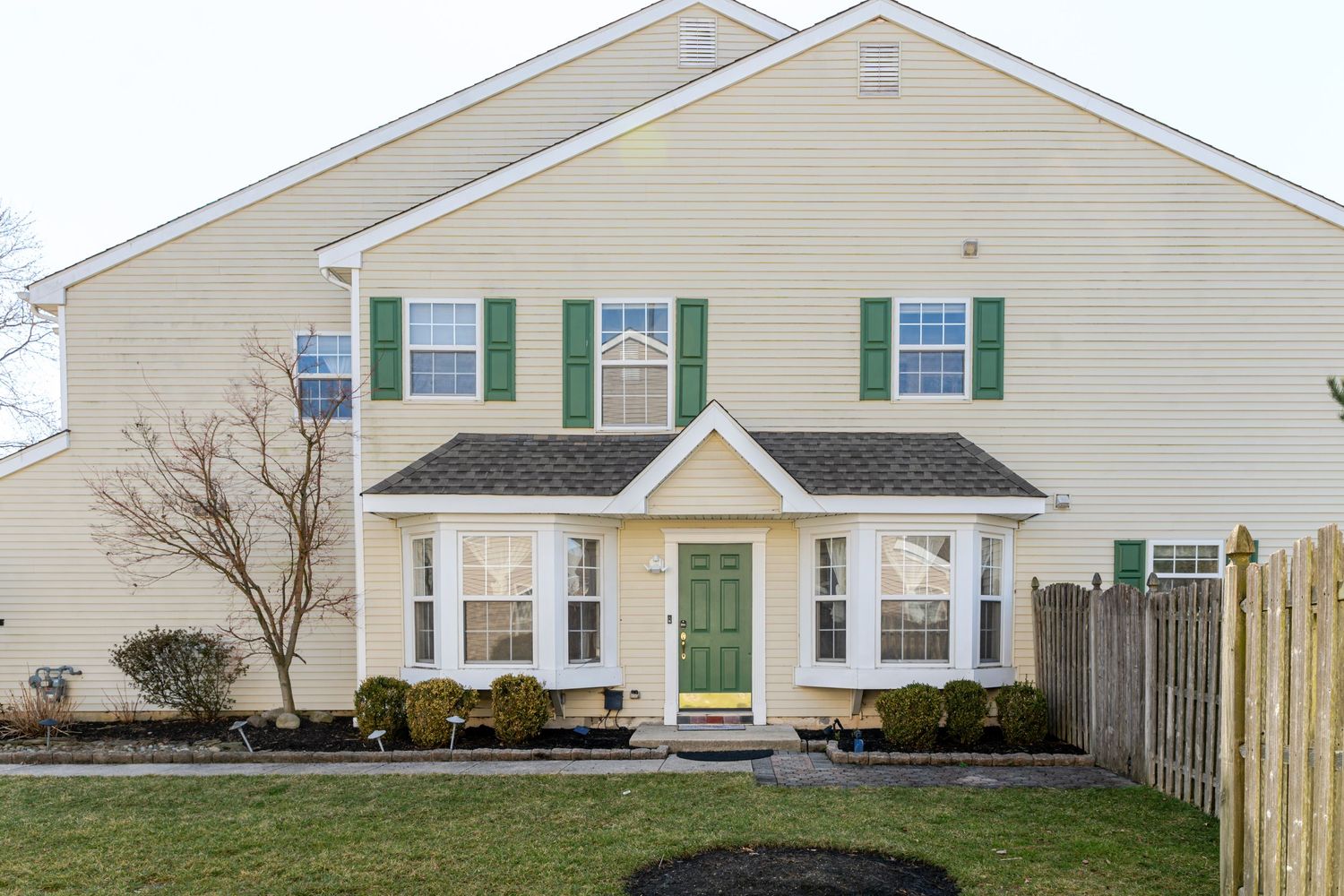 Two-story colonial home with green shutters, matching front door, beige siding, and manicured lawn on a cloudy day.