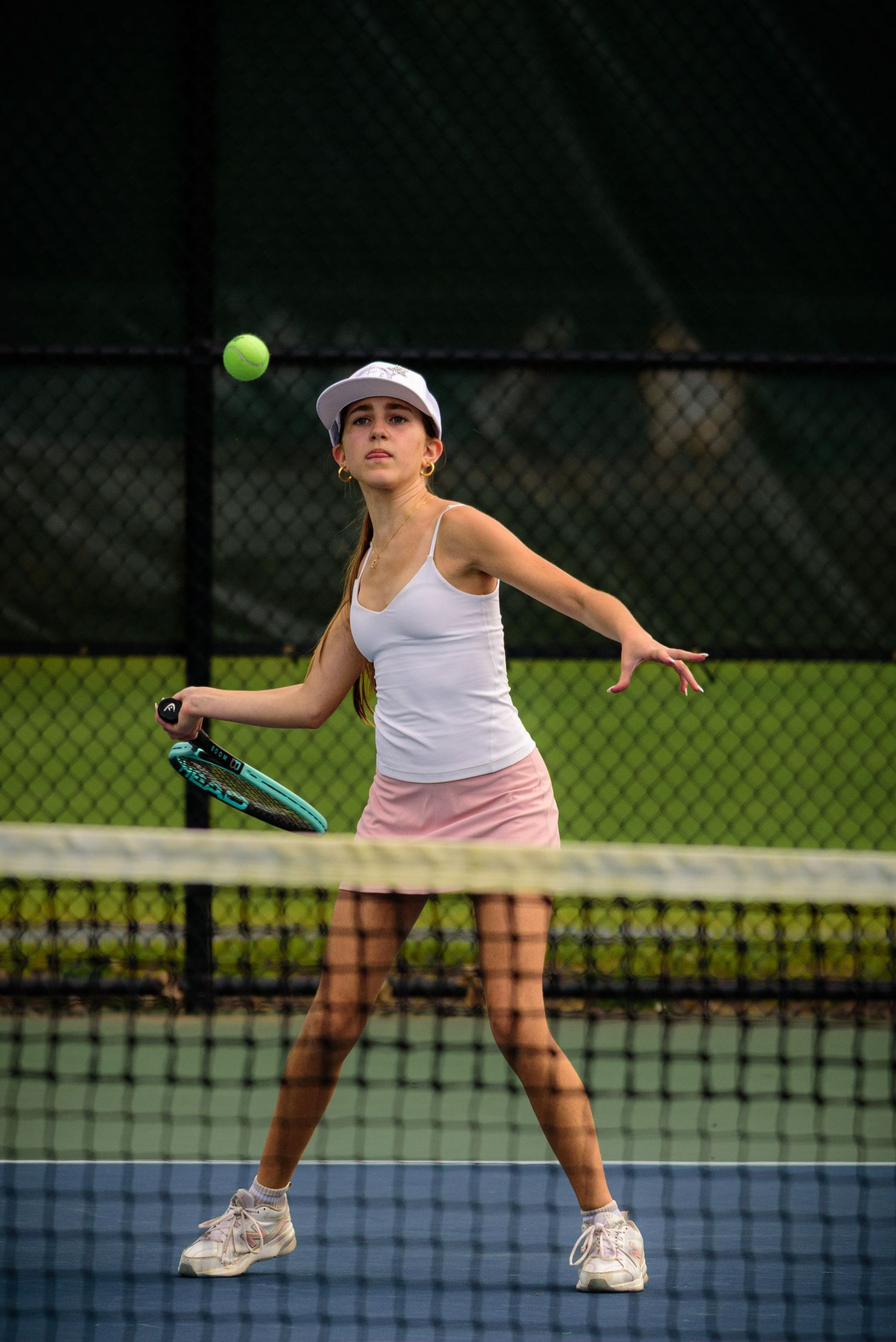 Tennis player in white tank top and pink skirt hits a forehand shot on outdoor tennis court.