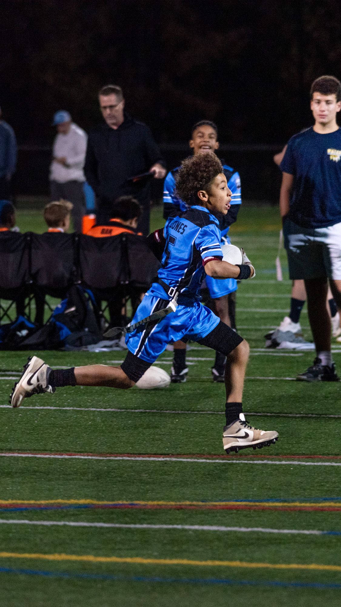 Young football player in blue uniform sprinting on turf field during a youth flag football game at night.