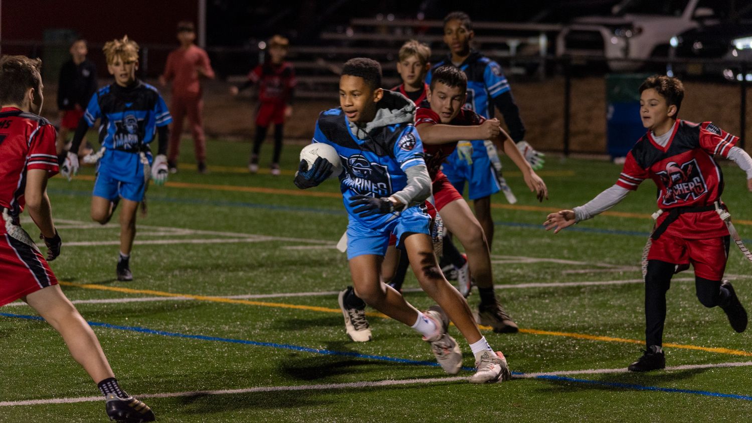 Youth flag football players compete under stadium lights, with a player in blue uniform running with the ball.