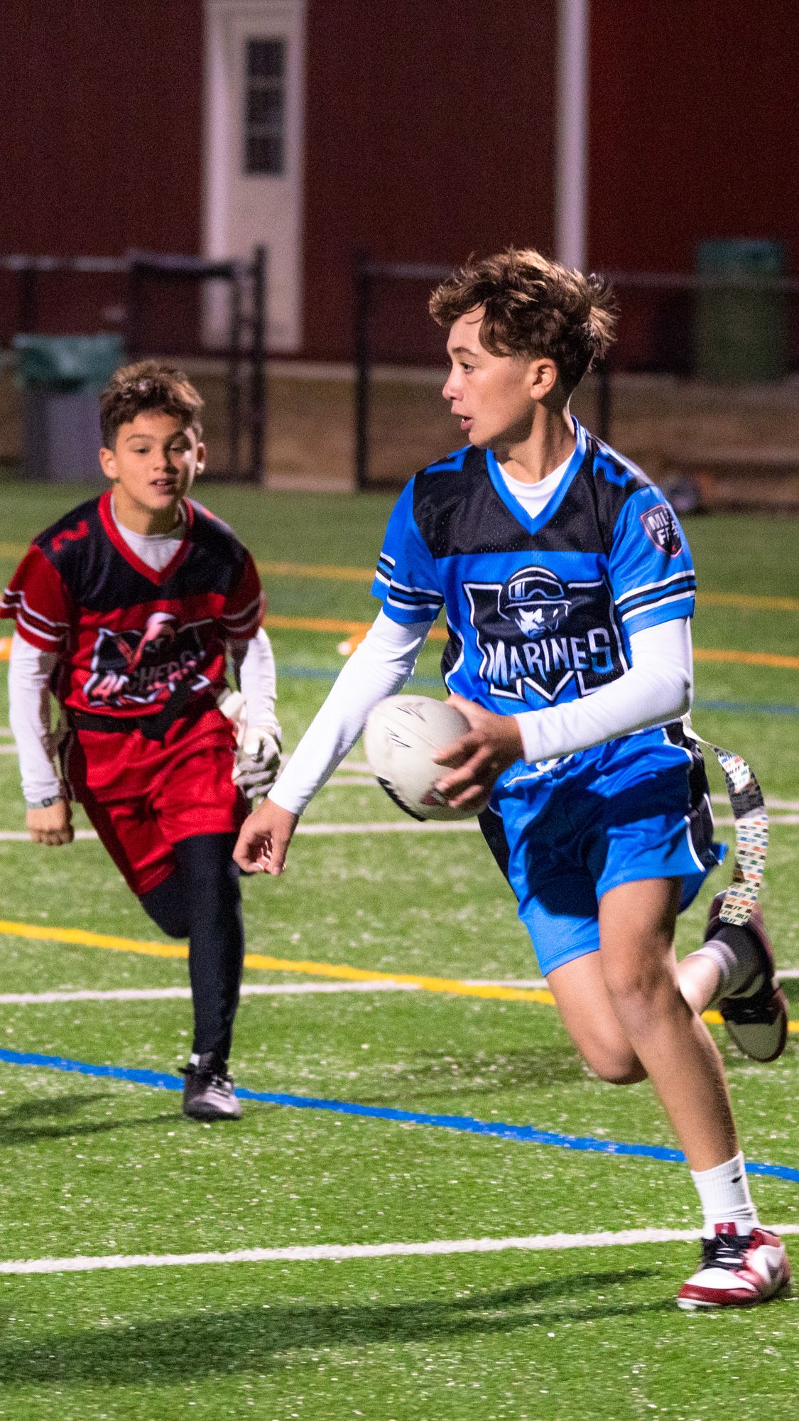 Two youth flag football players in action, Marines player in blue carrying ball, defender in red pursuing on green turf.