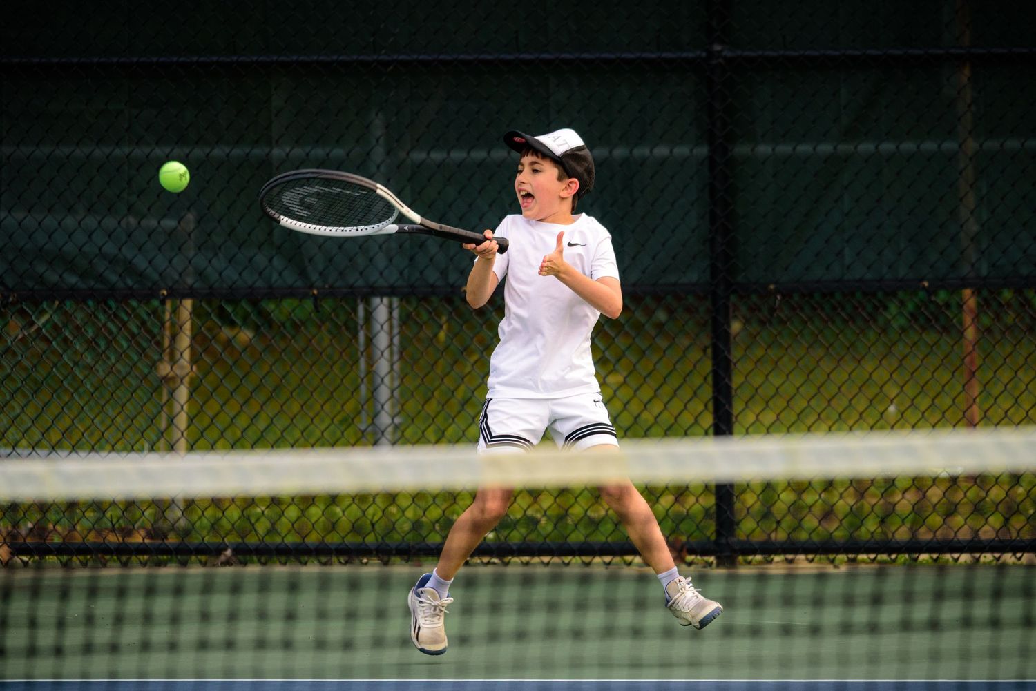 Tennis player in white attire hits a forehand shot on an outdoor tennis court during a match.