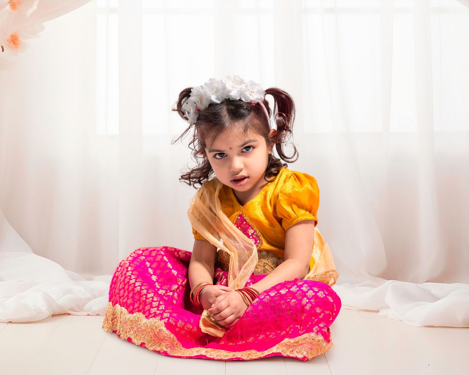 Young Indian girl in traditional pink and yellow lehenga with flower hair accessory sits gracefully on white floor.