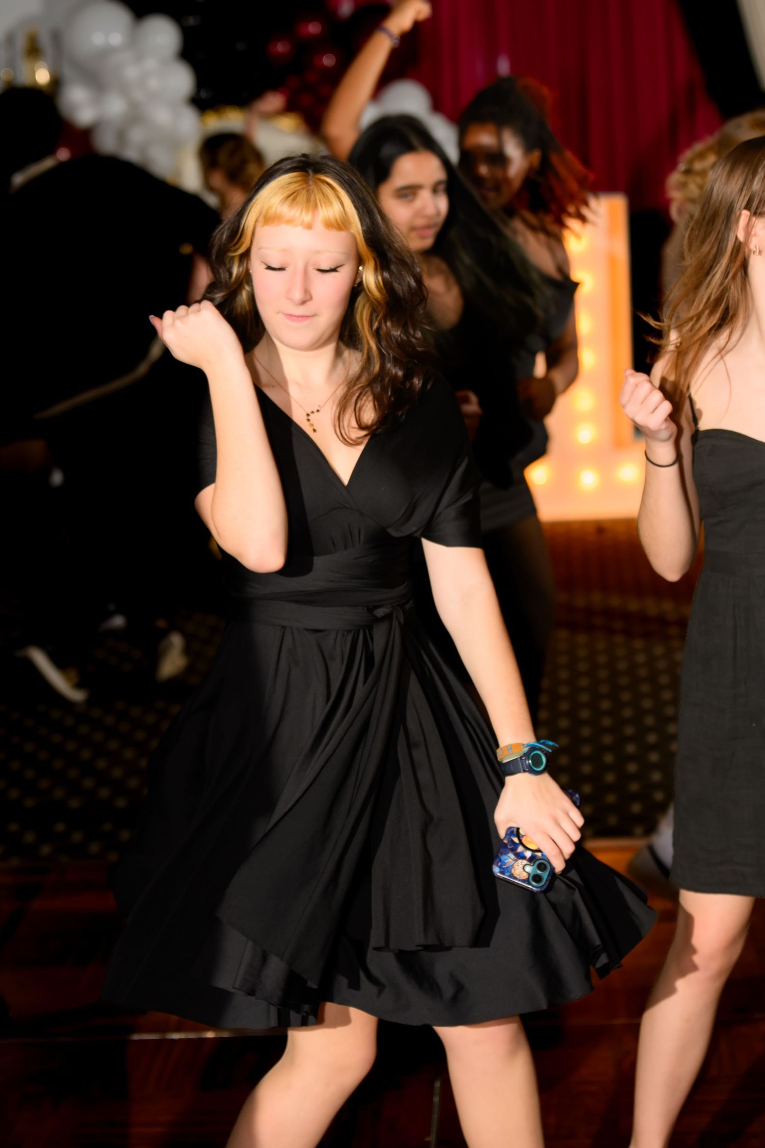 Young woman in black dress dancing at a formal party event with other guests and balloon decorations.