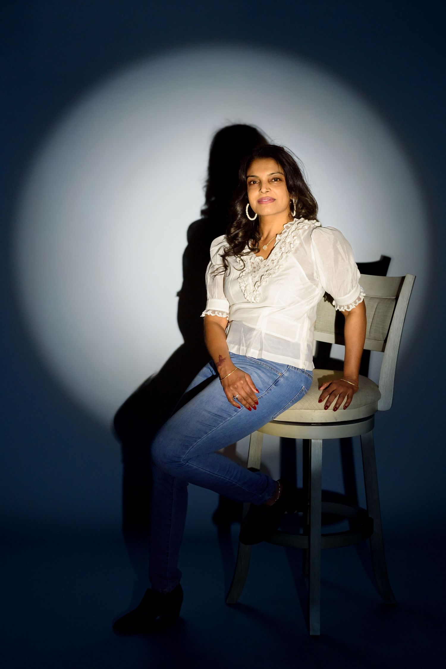 Woman in white blouse and jeans sits on white chair under spotlight with dramatic shadow on blue background.