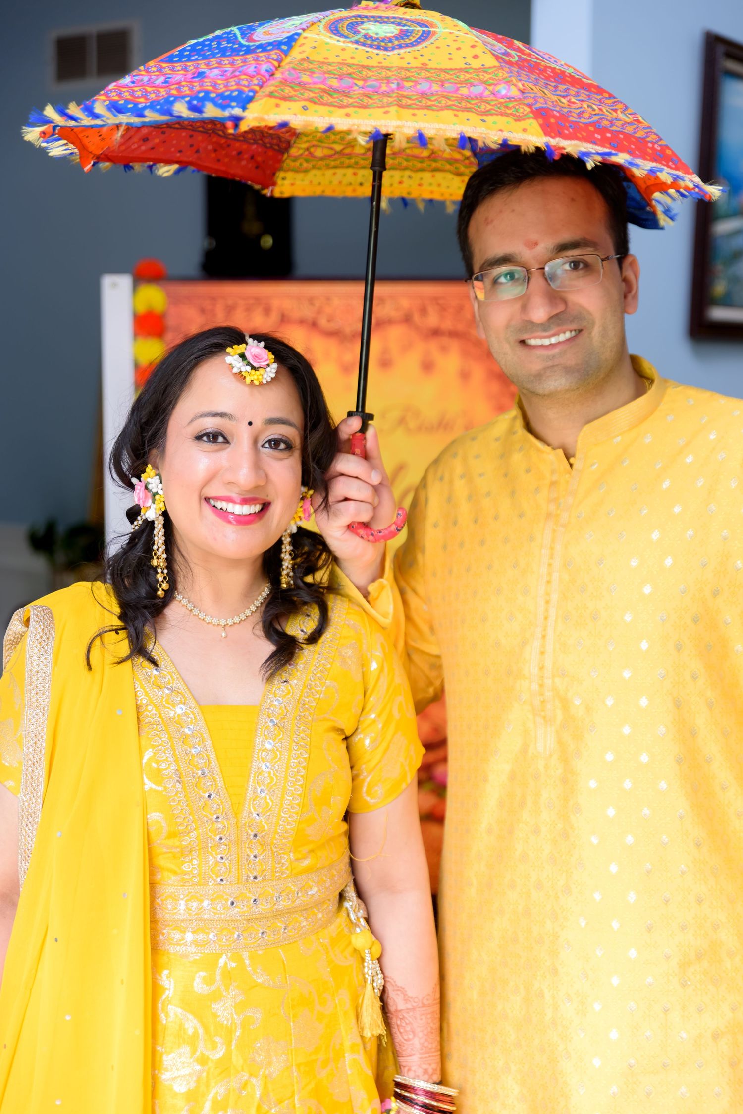 A smiling Indian couple in matching yellow traditional outfits poses under a colorful decorative umbrella during a festive celebration.