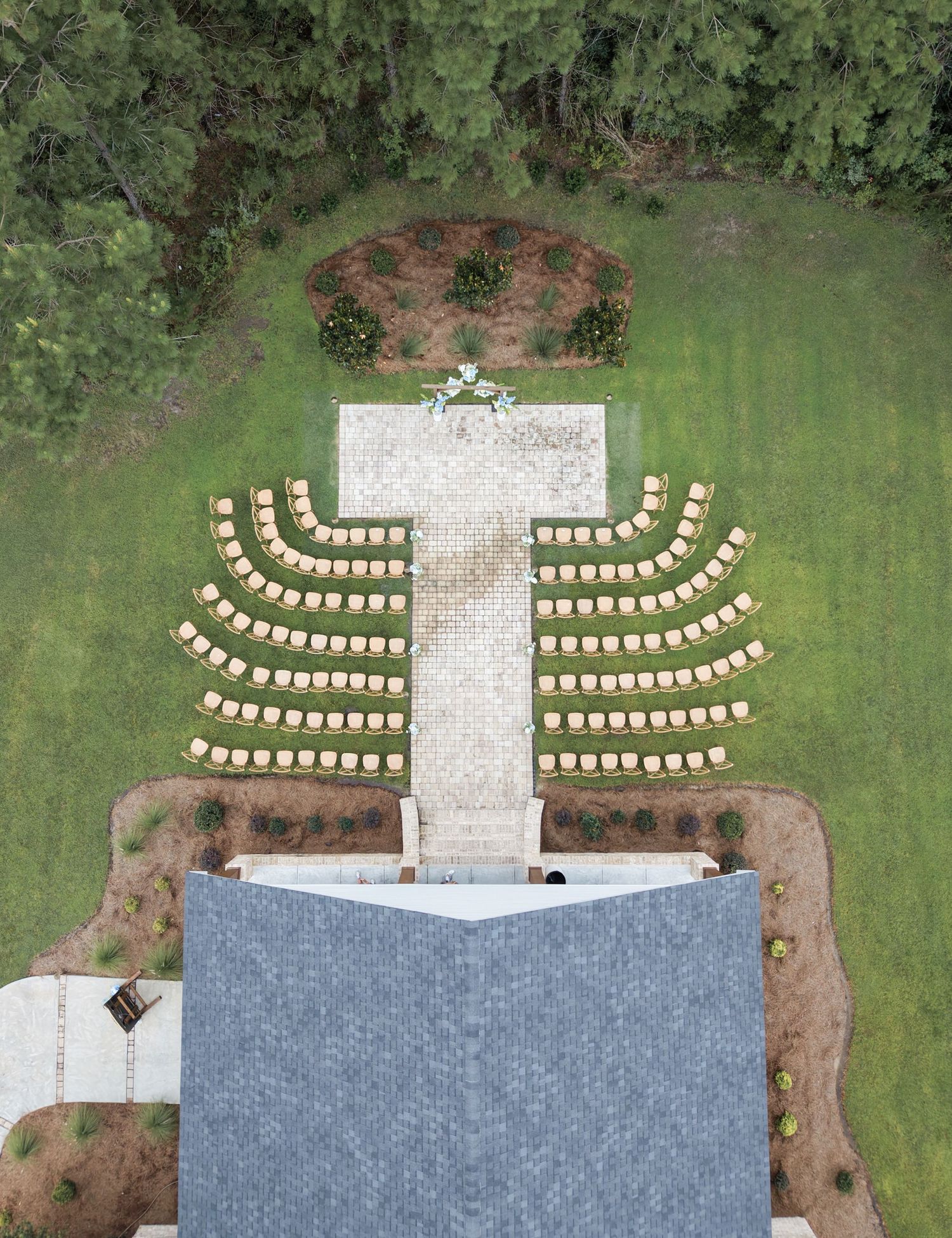 Aerial drone view of an outdoor wedding ceremony setup arranged in a cross shape on a green lawn.