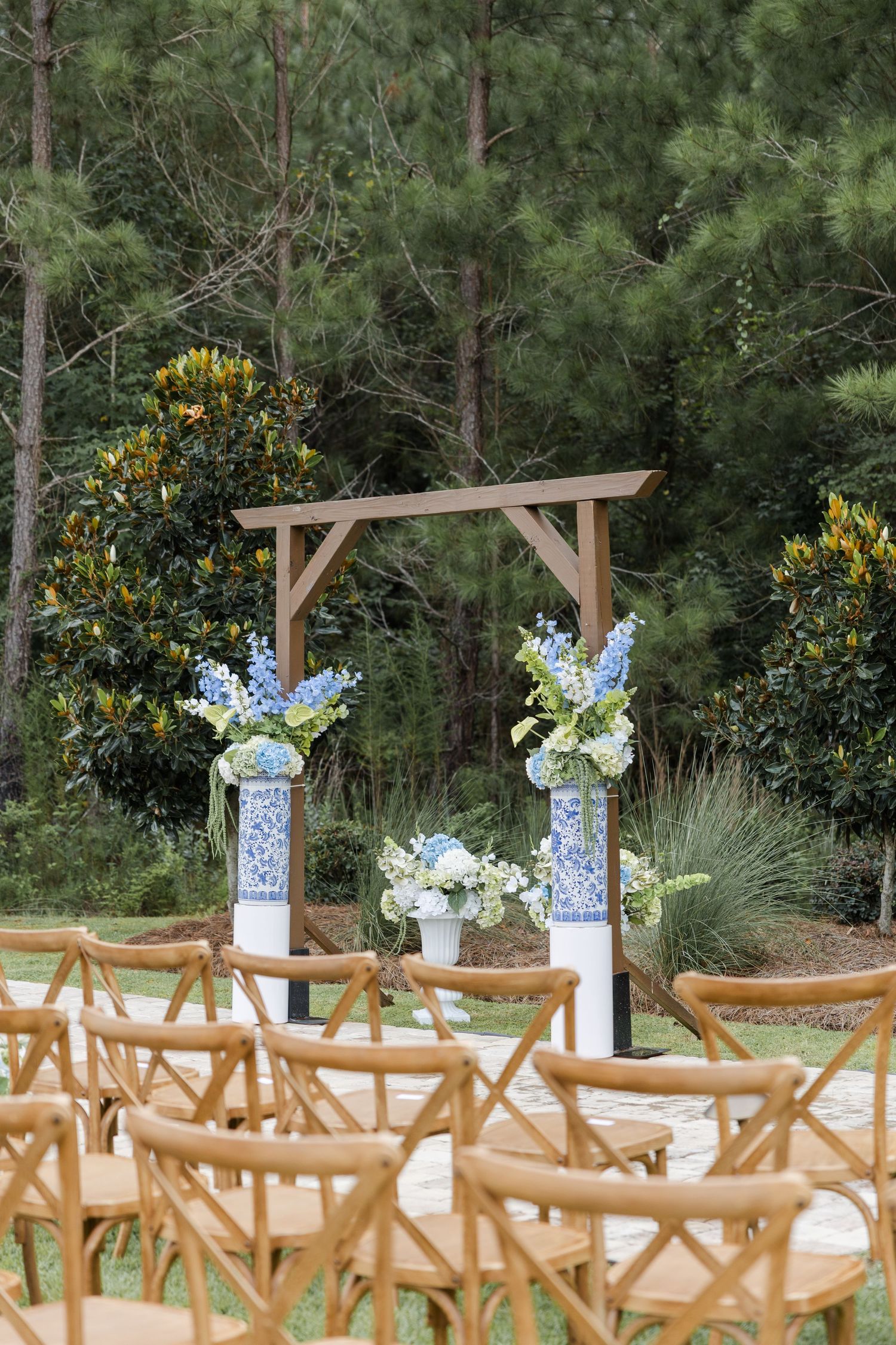 Outdoor wedding ceremony arch with blue and white floral arrangements and wooden cross-back chairs.