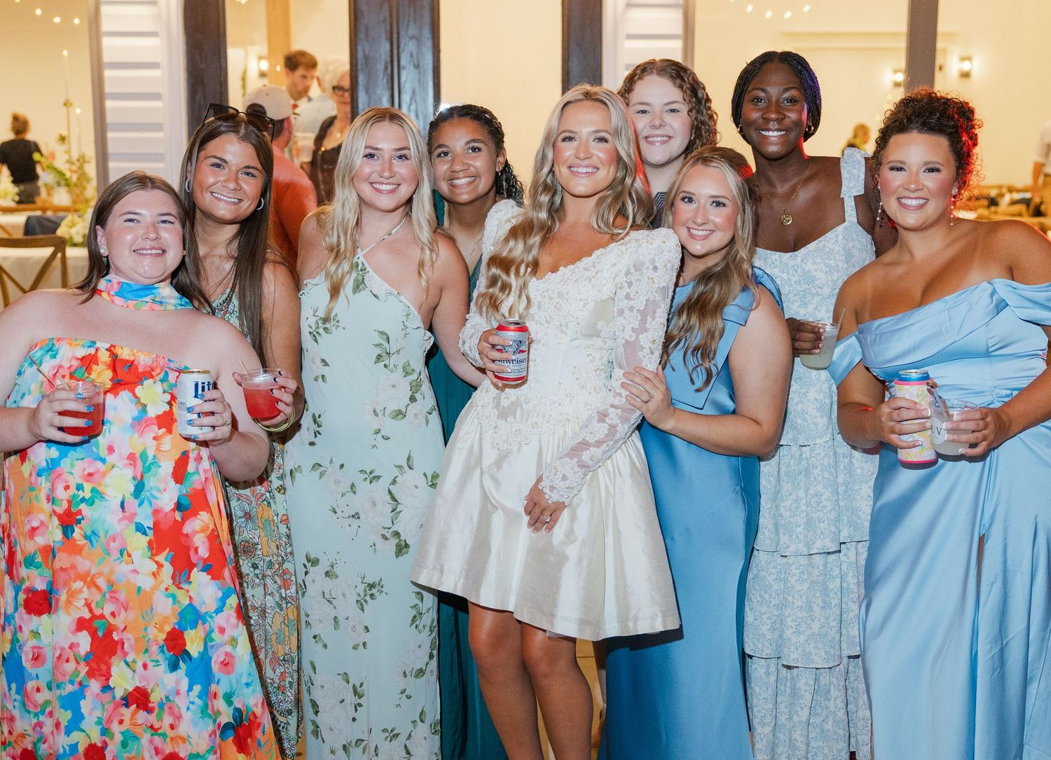 Bride in short white reception dress posing with bridesmaids and guests in colorful and blue dresses at wedding reception.
