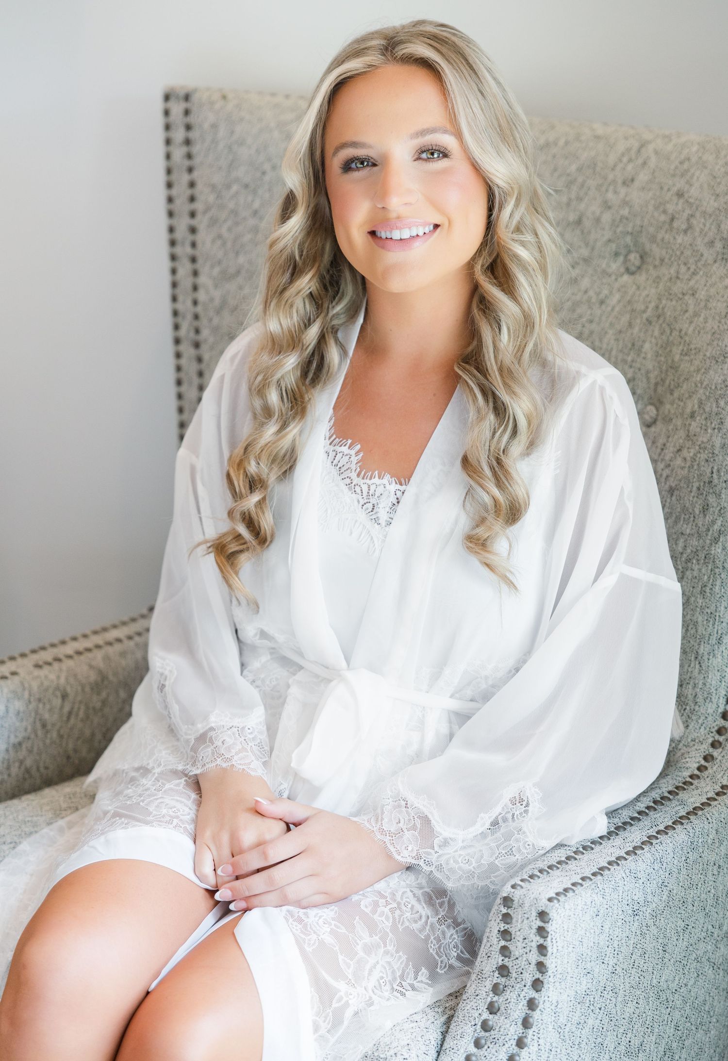 Smiling bride with long blonde waves wearing a white lace robe seated in a gray tufted chair.