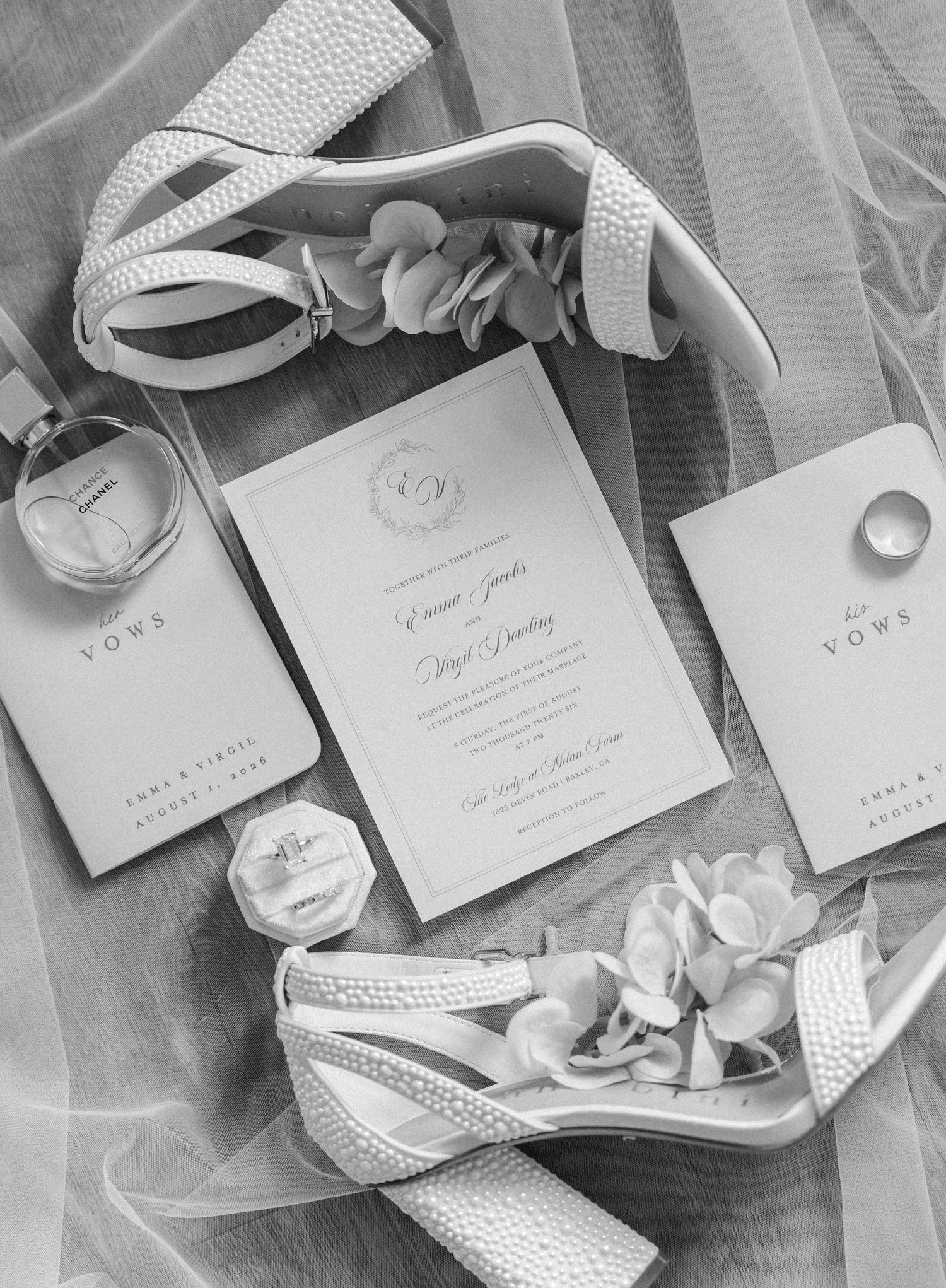 Black and white flat lay of wedding invitation suite with pearl heels, vow books, ring box, and perfume on sheer veil.