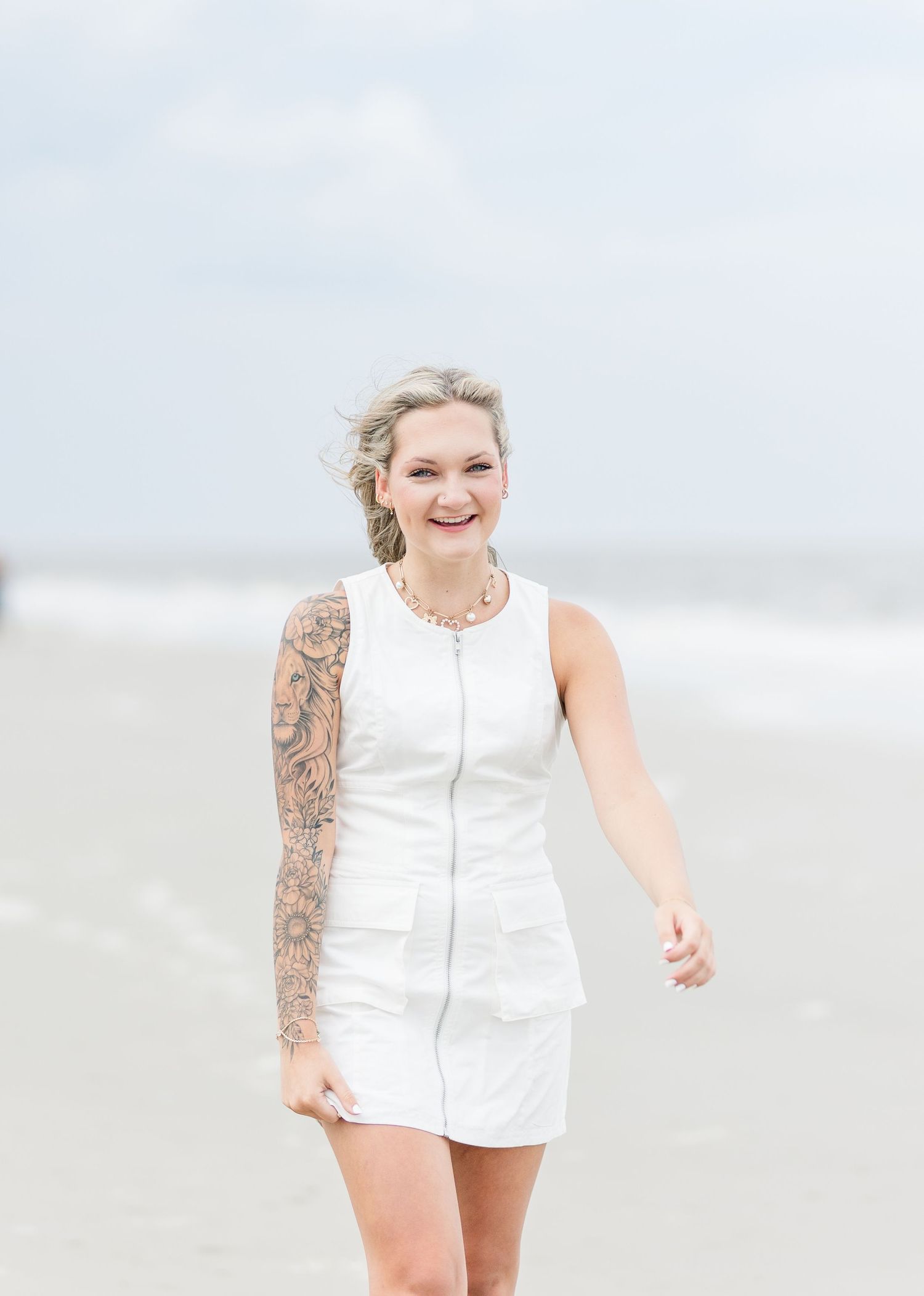 A person in a white sleeveless dress poses on a beach with waves visible in the background.