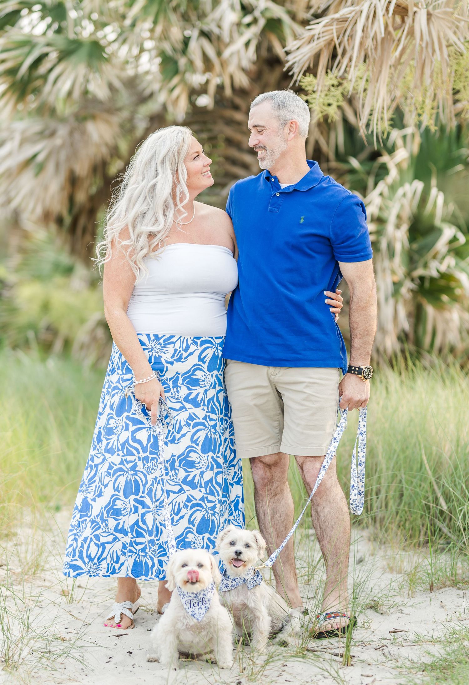 Couple in blue and white outfits walks their small white dog along a beach path surrounded by palm trees.