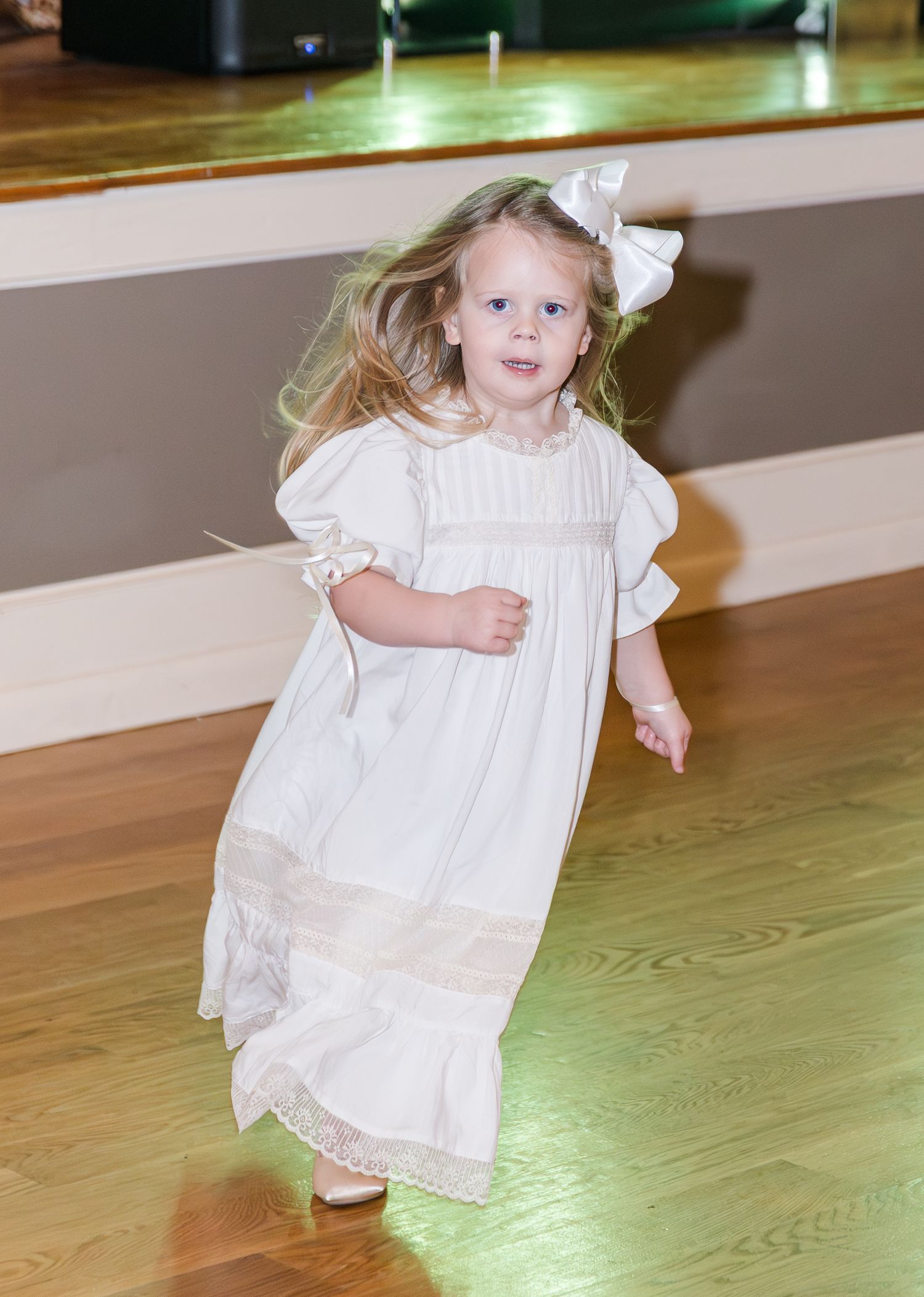 Adorable flower girl in white dress with bow running on dance floor at a wedding reception.