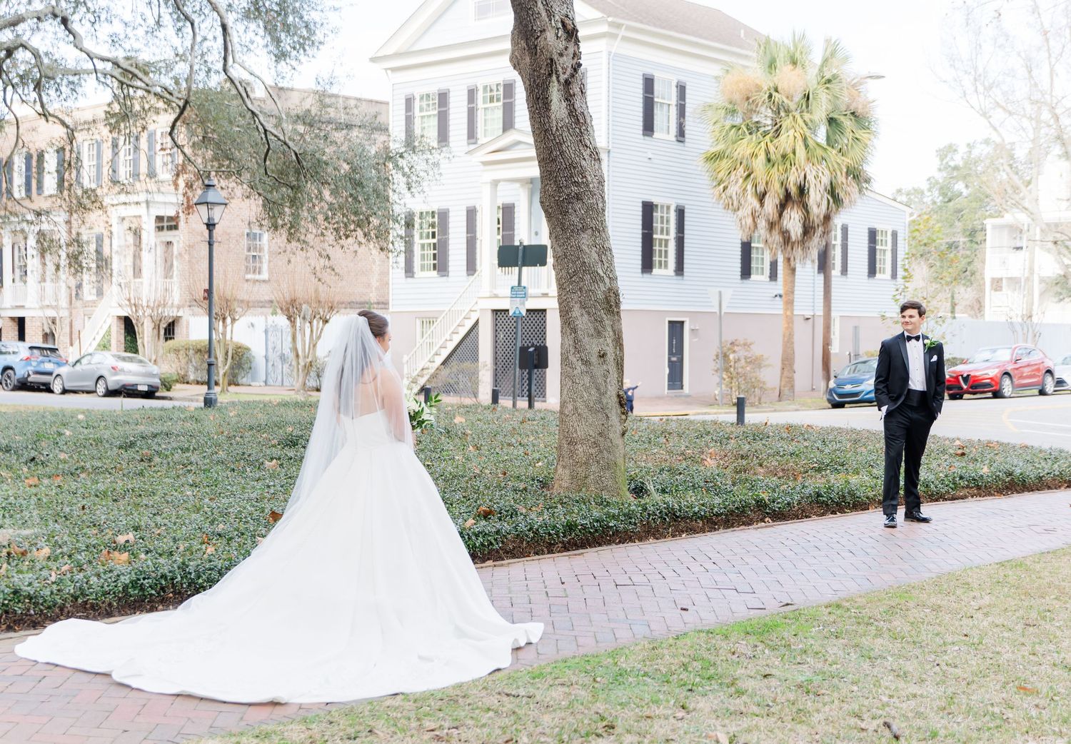 Bride in long white gown and veil approaches groom for first look on brick path at outdoor wedding.