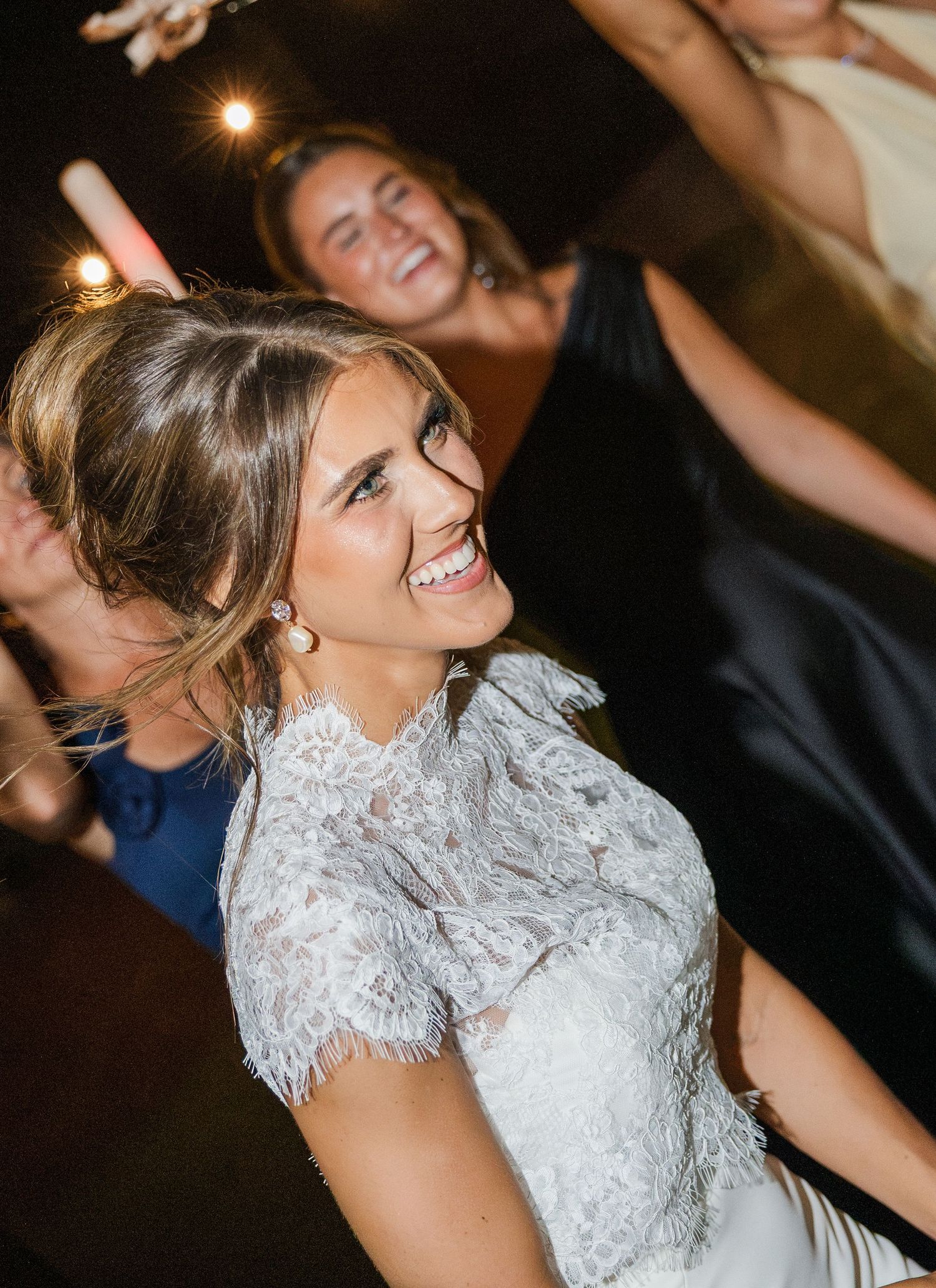A joyful bride in a white lace dress laughs and dances with friends during her wedding reception celebration.