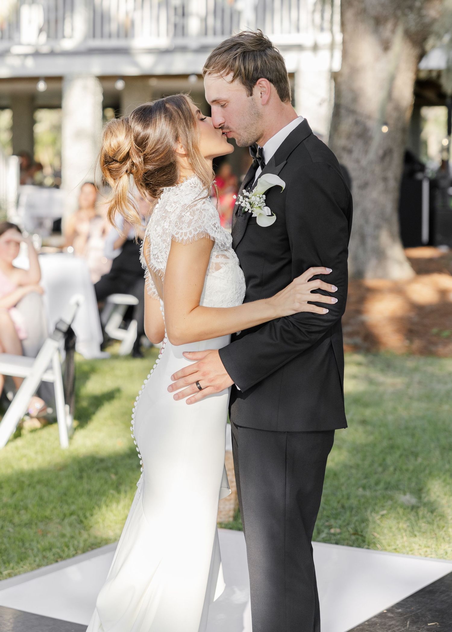 Bride and groom share first kiss in elegant wedding attire outdoors, she in lace gown, he in black tuxedo.
