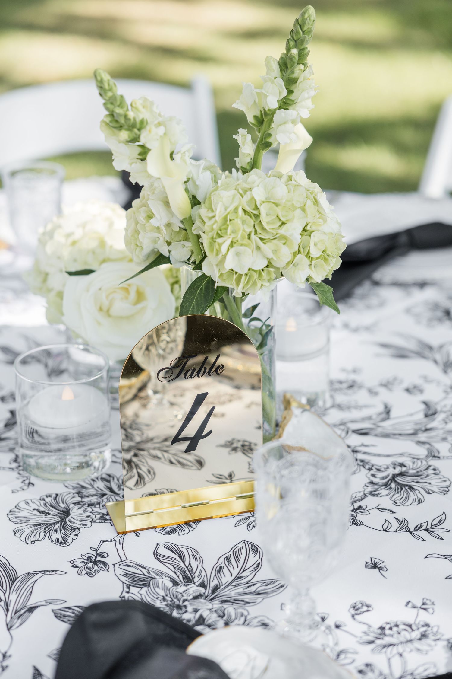 Elegant wedding table 4 centerpiece with white hydrangeas, snapdragons, gold mirror number sign on black and white floral tablecloth.