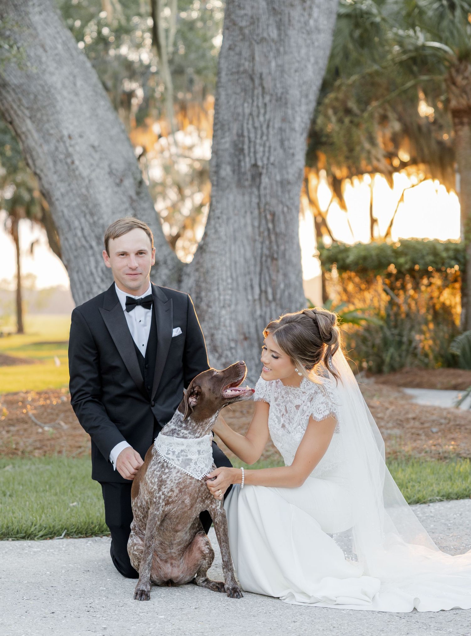 Bride and groom pose with their spotted dog wearing a white collar on their wedding day outdoors.
