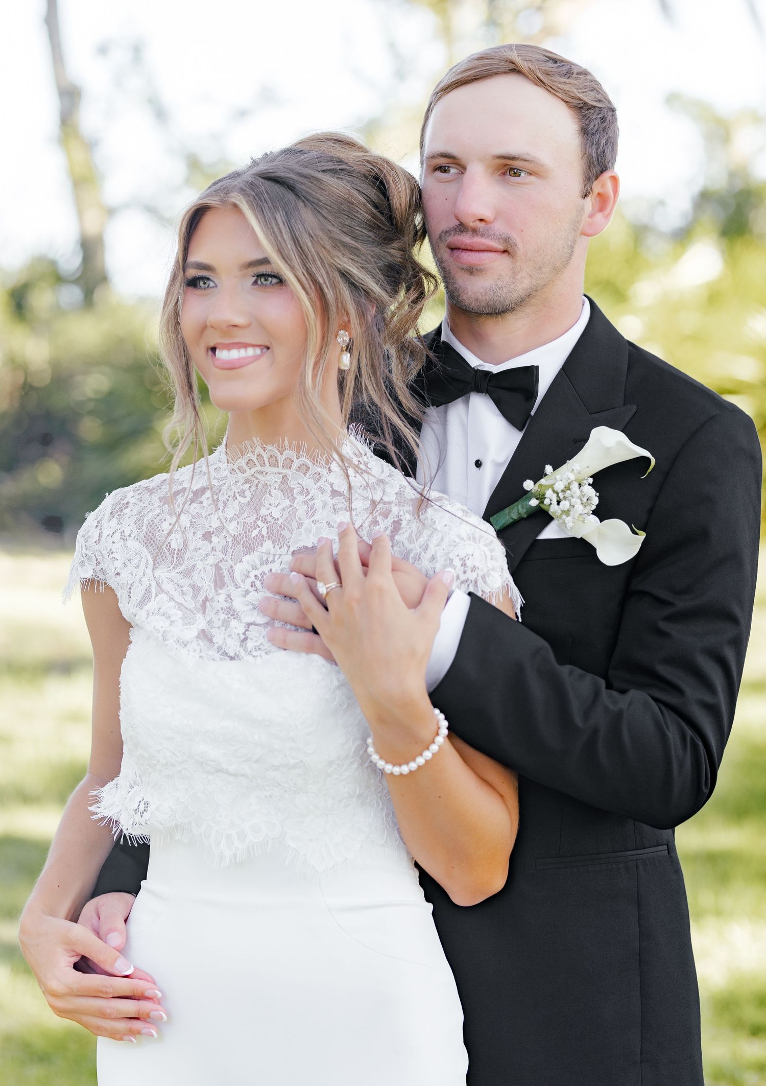 Bride in lace cap sleeve wedding gown and groom in black tuxedo with white calla lily boutonniere pose outdoors.