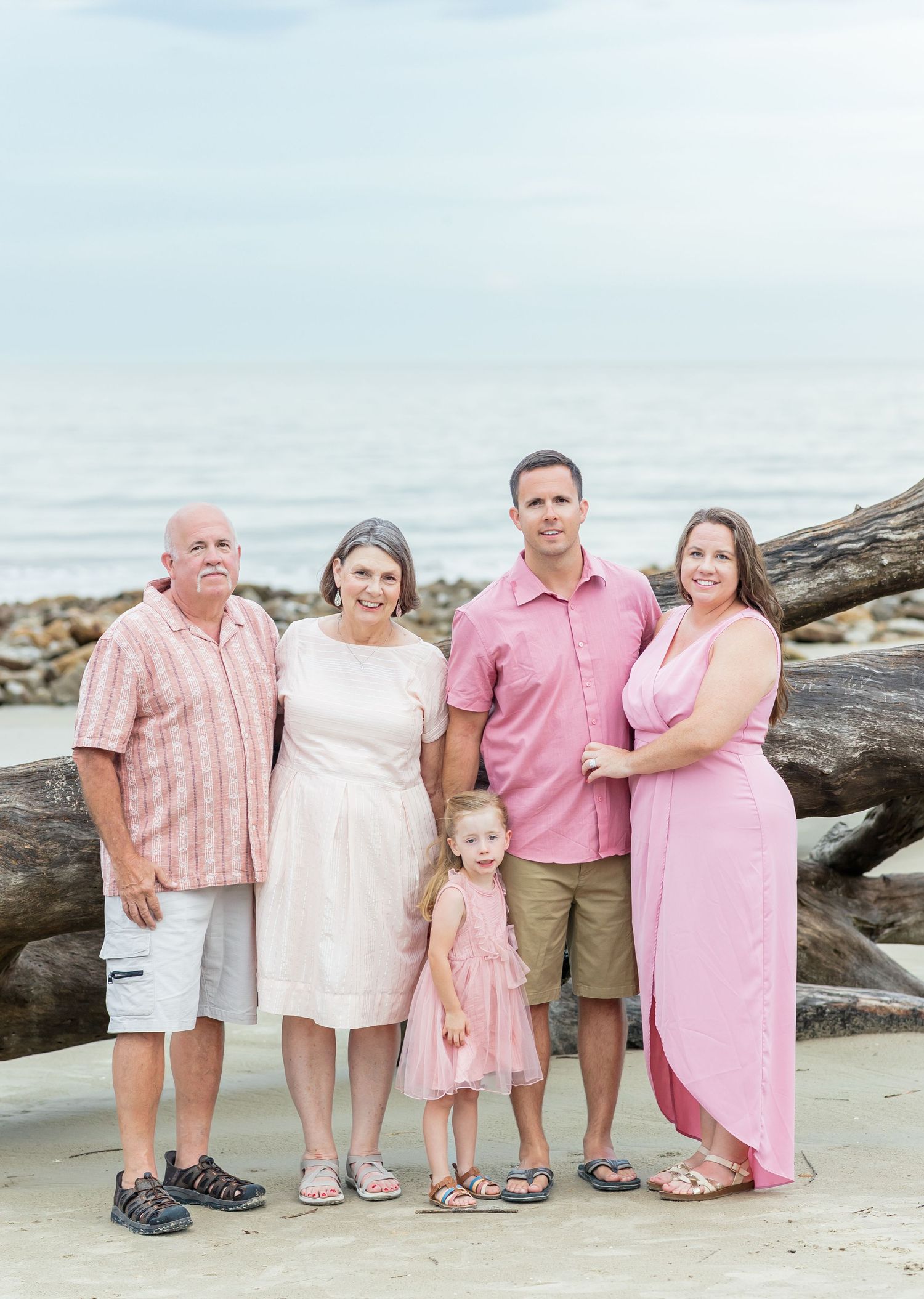 Family portrait session at the beach with coordinating pink outfits against coastal rocks and water backdrop.