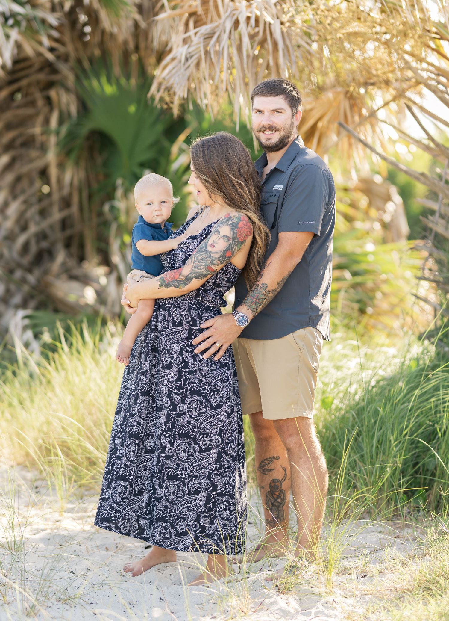 A family poses together on a beach with palm trees, wearing casual summer attire including a floral maxi dress and khaki shorts.