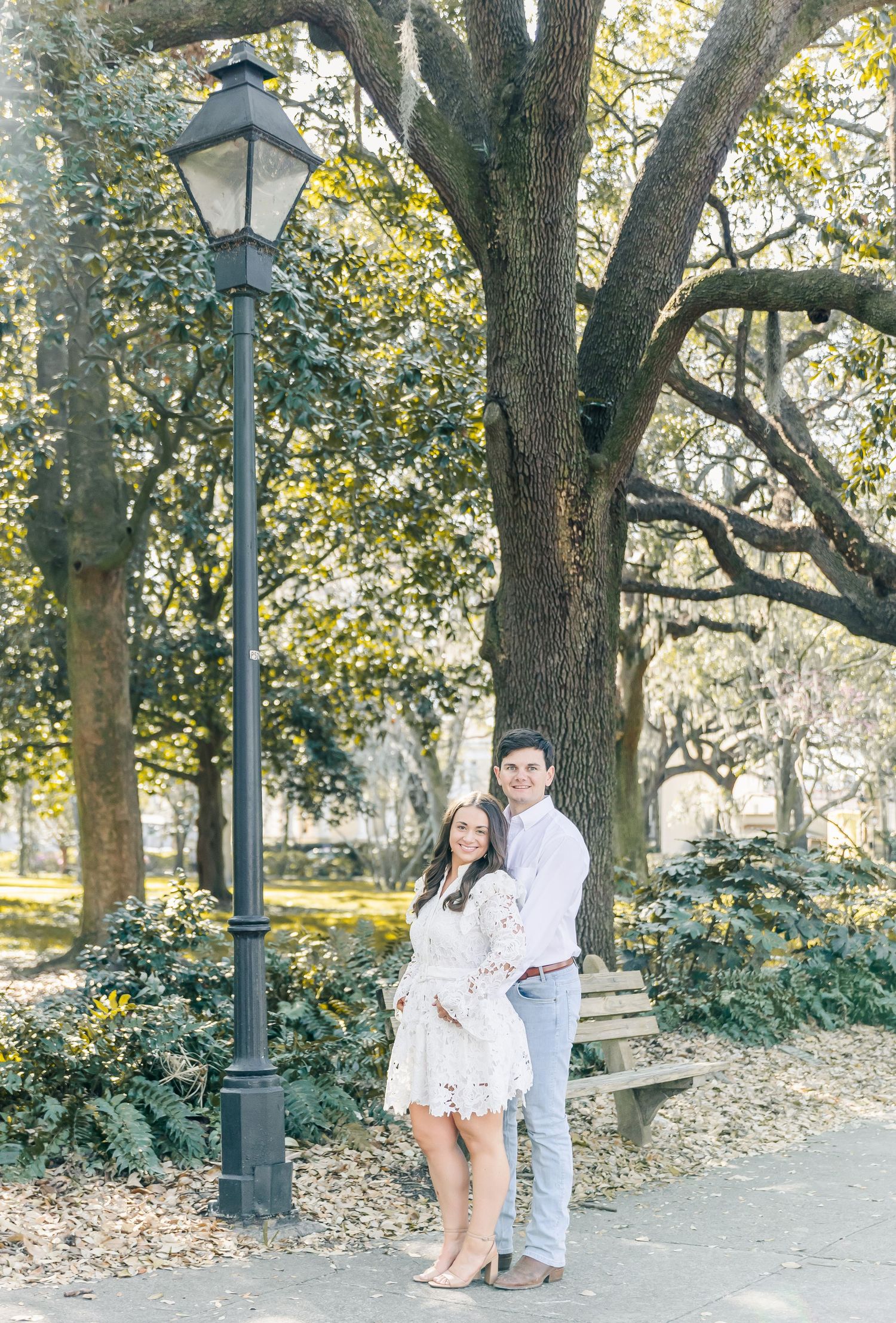 A couple poses together beneath a historic lamppost and oak tree in a romantic garden setting.